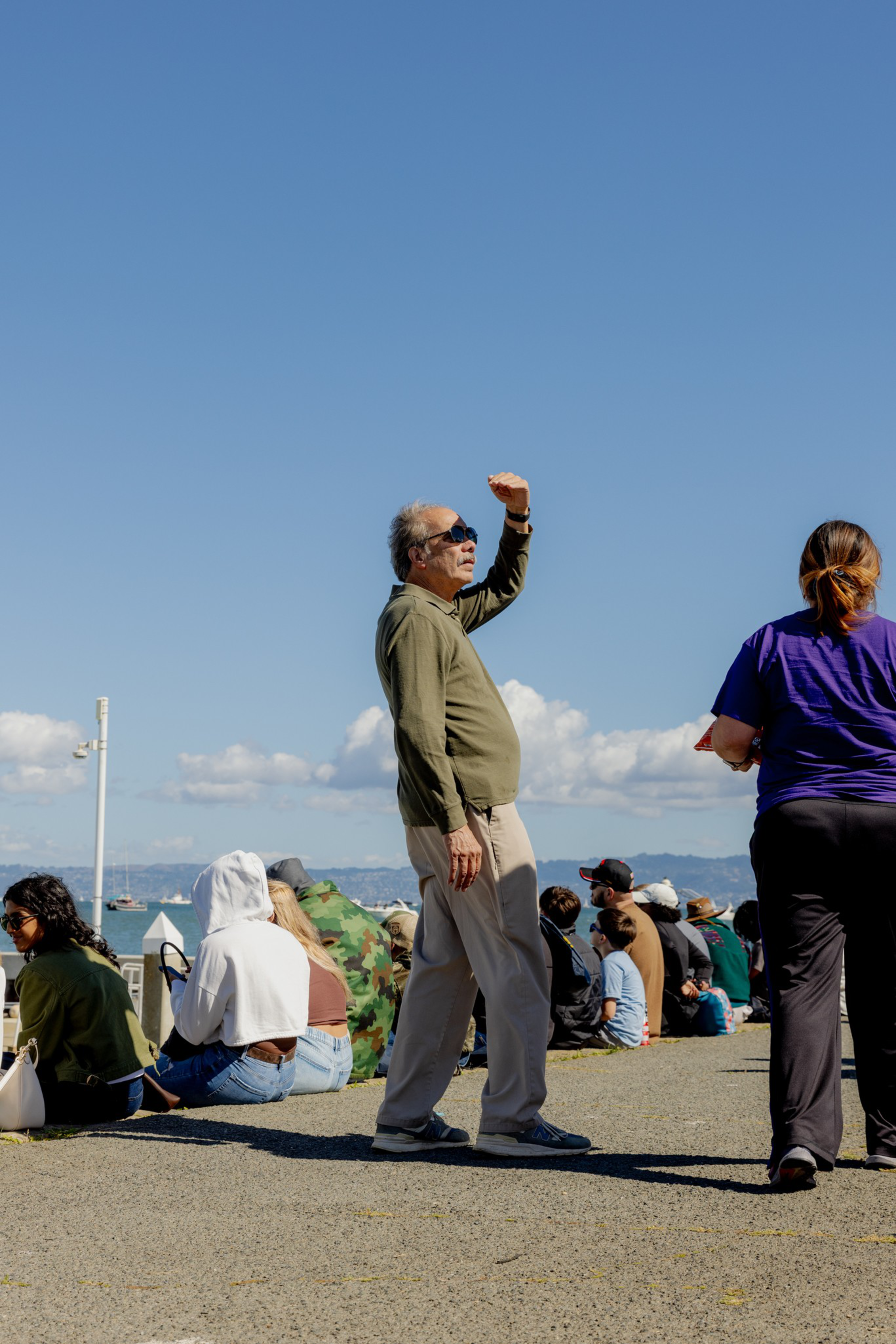 A man in sunglasses and light pants shields his eyes from the sun while standing near a group of seated people by the water on a clear day.
