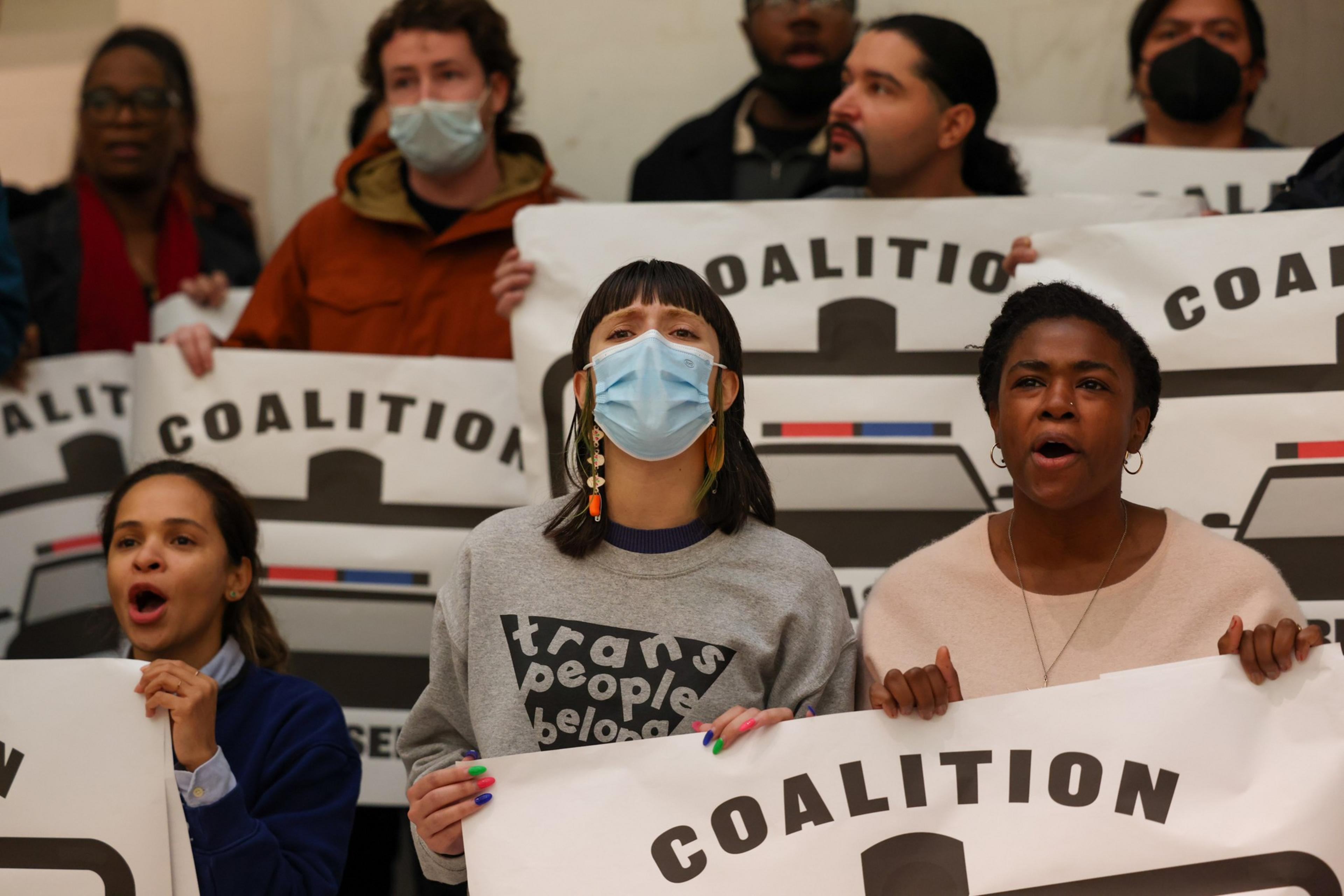 A diverse group of people passionately hold signs that say "COALITION," with one person wearing a mask and a shirt reading "trans people belong."