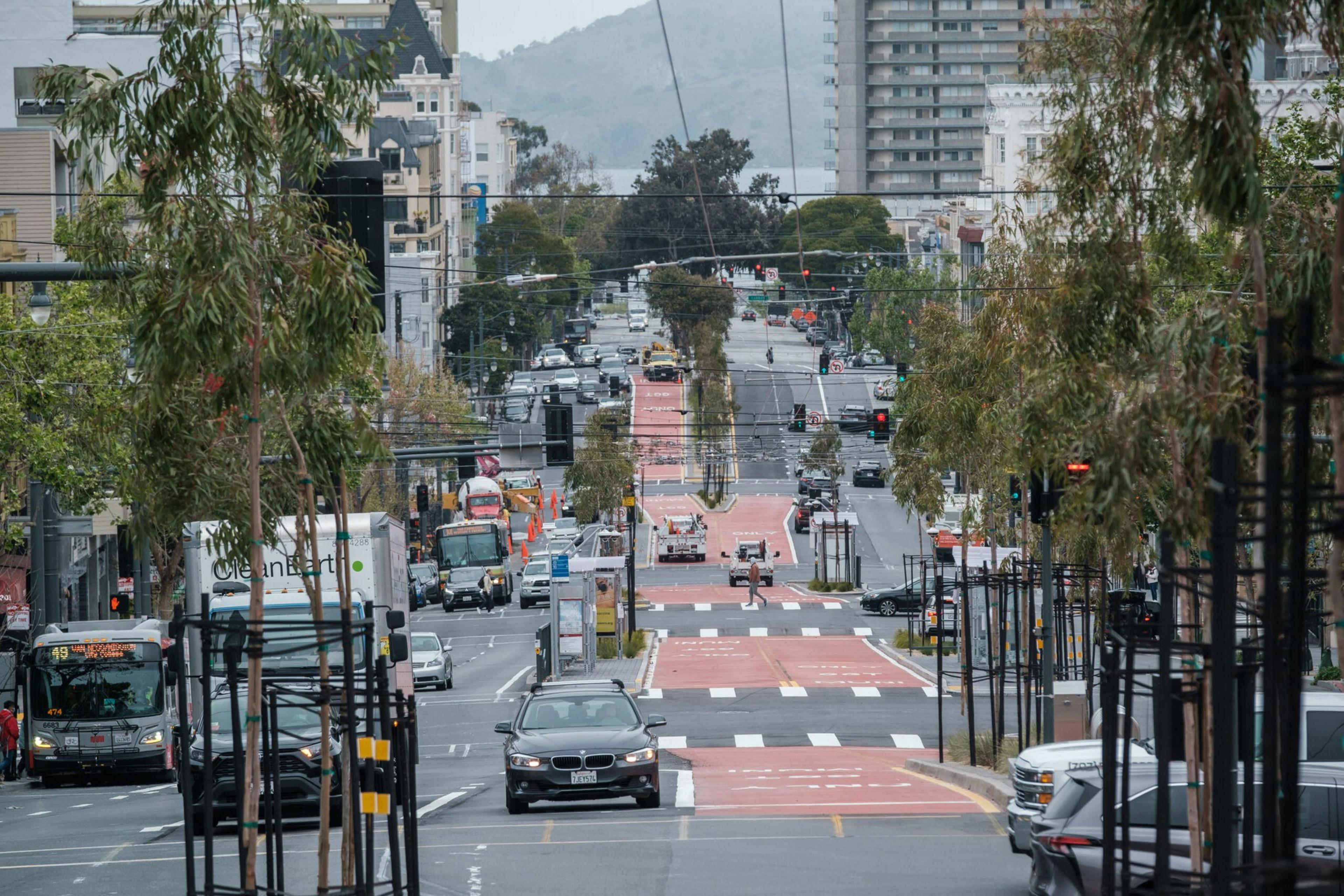 A busy urban street with cars, buses, and trucks travels uphill, flanked by trees and buildings on both sides, featuring multiple traffic lights and red bus lanes.