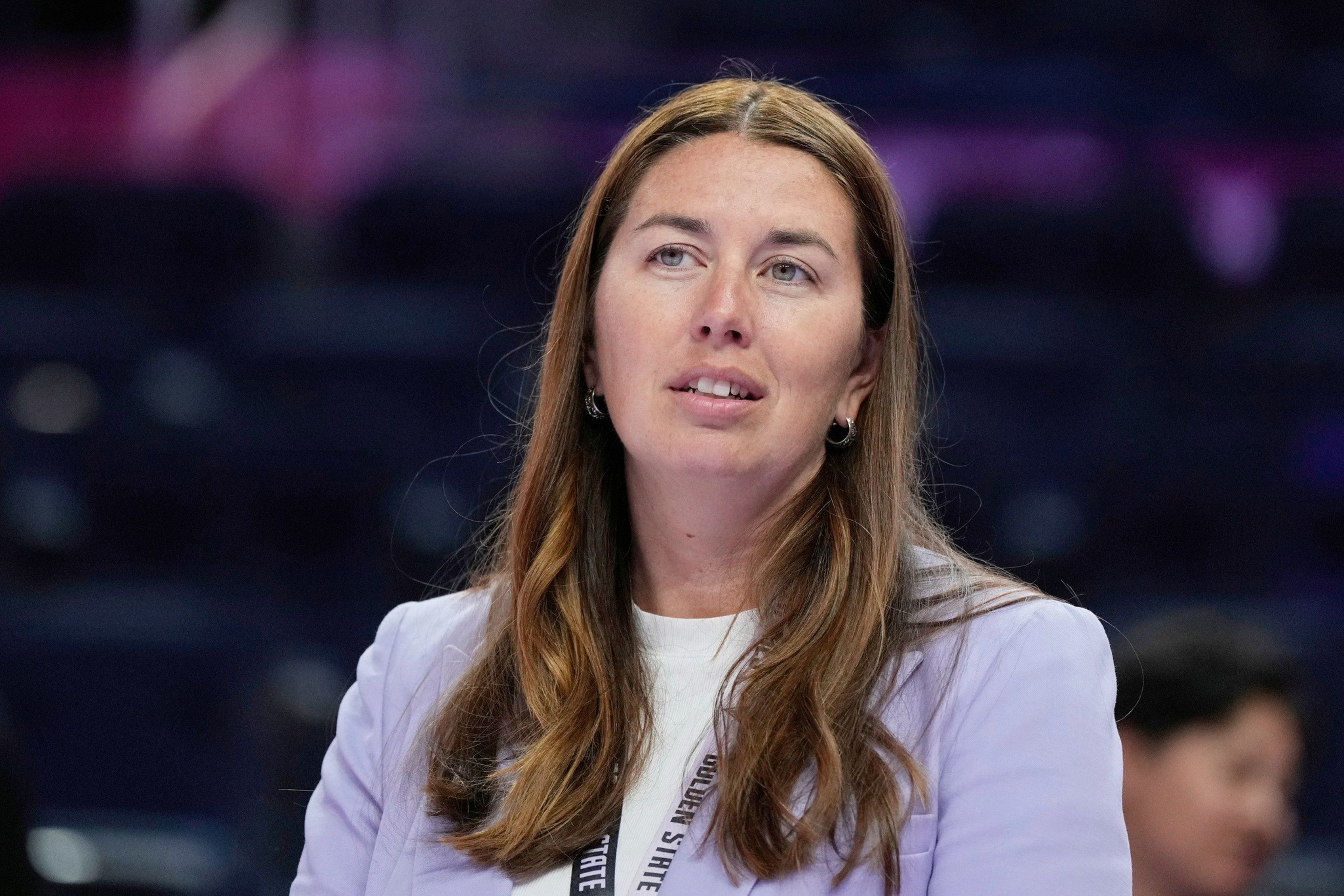 A woman with long brown hair wearing a light purple blazer and white shirt looks off to the side with a thoughtful expression.