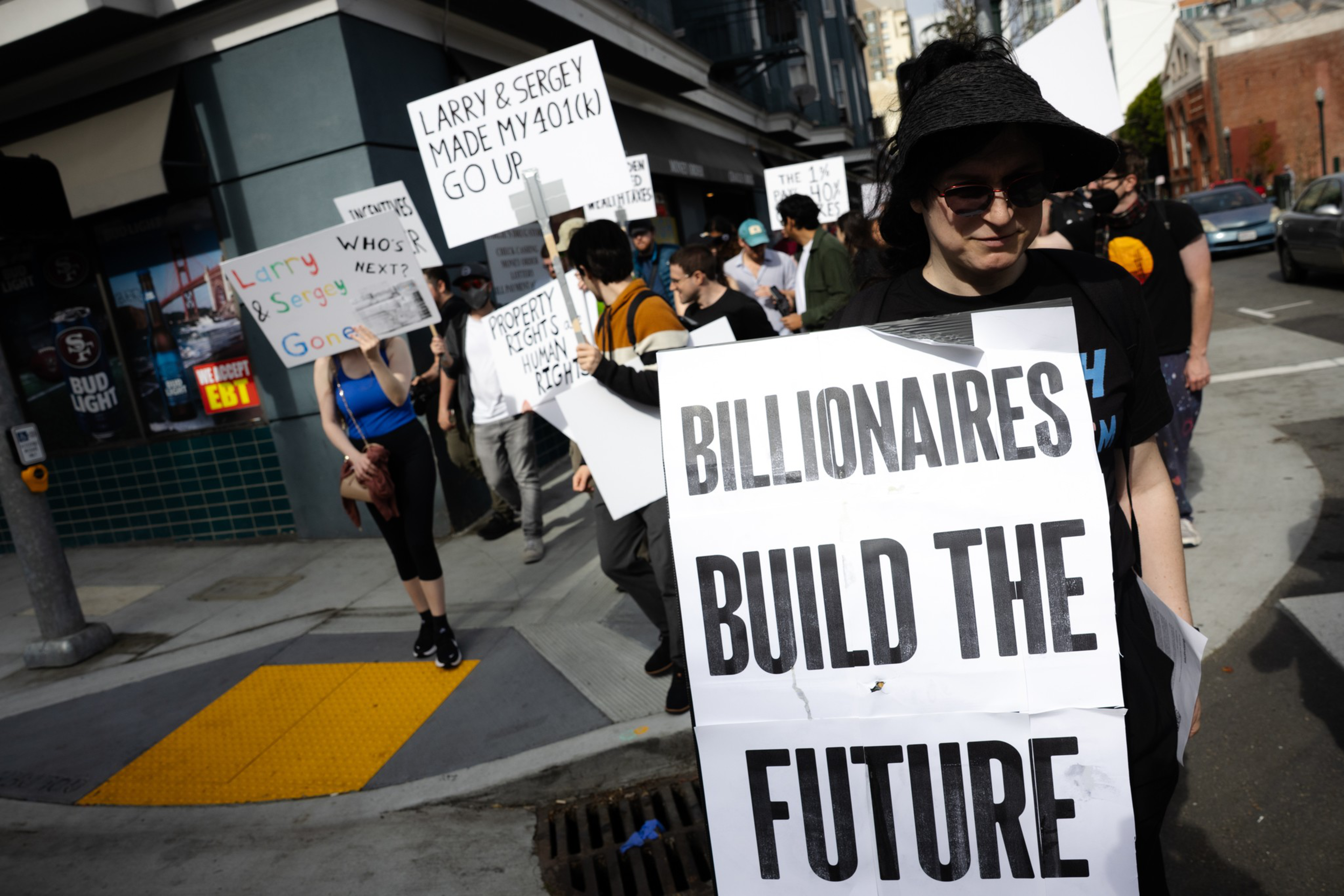 People march in a street protest holding signs with messages like "BILLIONAIRES BUILD THE FUTURE" and "LARRY & SERGEY MADE MY 401(K) GO UP."