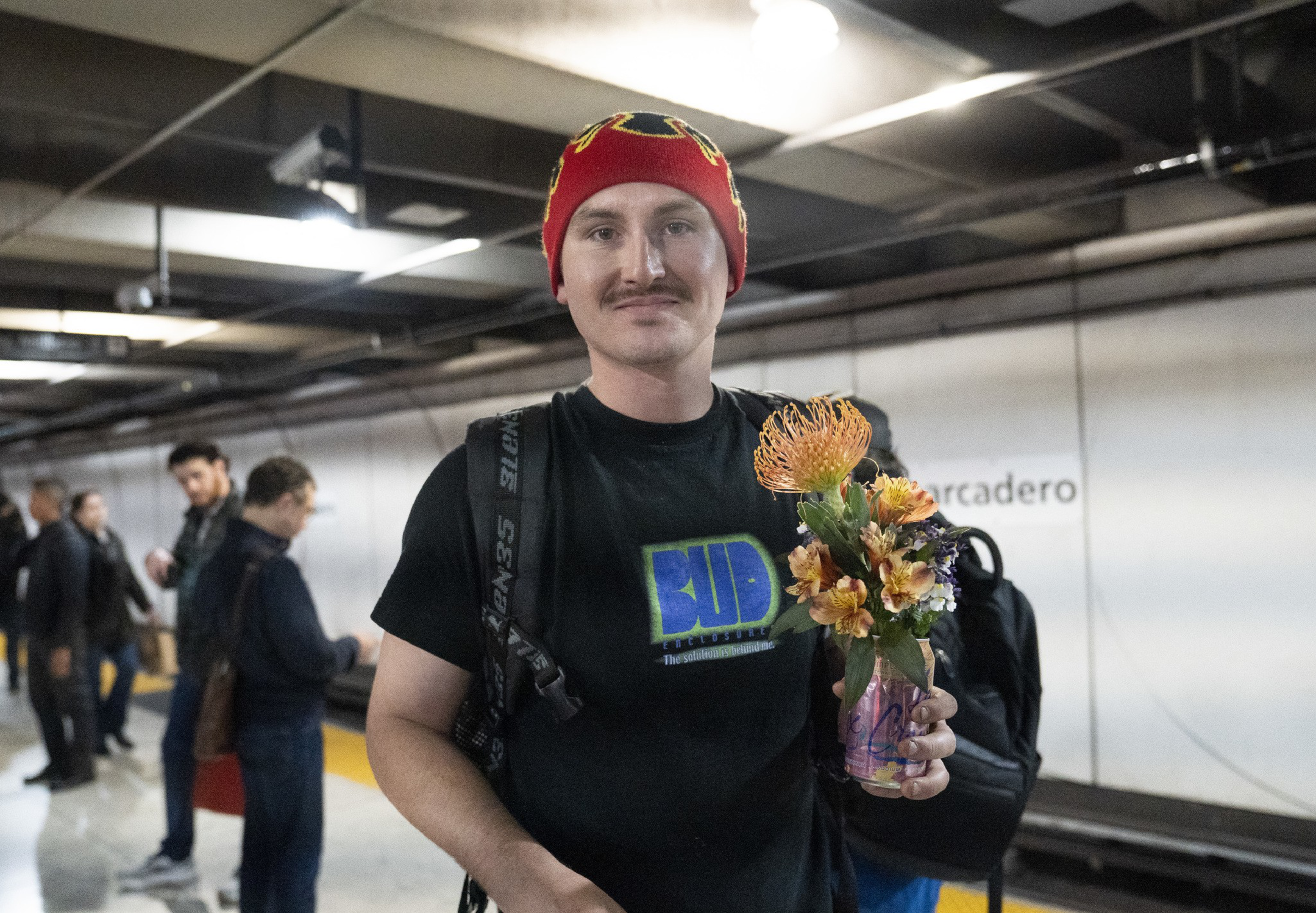 A person in a red beanie and black t-shirt holds a colorful bouquet in a can, standing on a subway platform with others in the background.