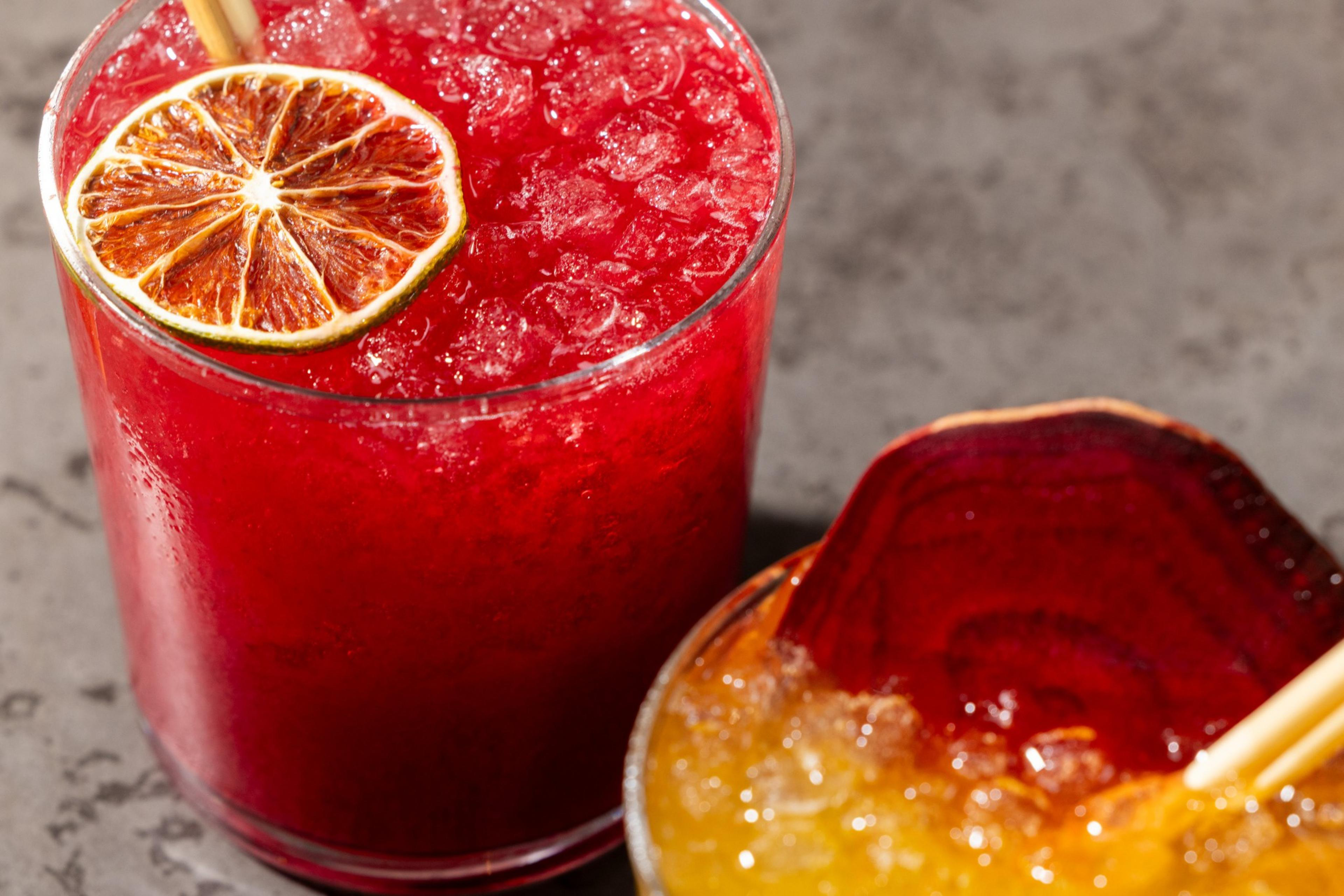 A vibrant red drink with crushed ice and a dried citrus slice sits beside an orange-yellow drink with a beet garnish, both in clear glasses on a gray surface.