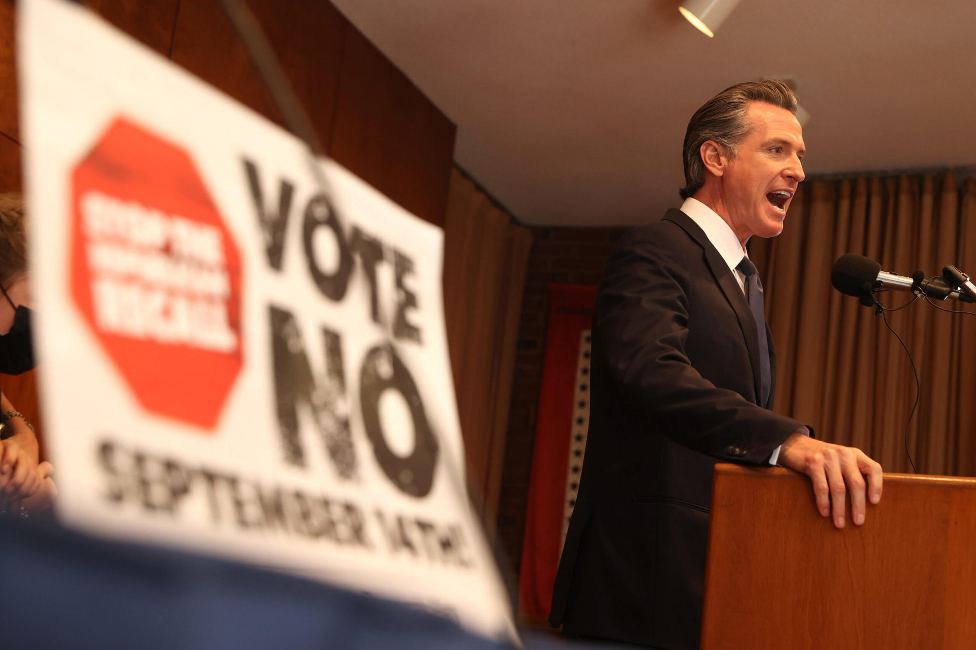 A man in a suit speaks at a podium with a microphone, while a blurred sign in the foreground reads "Vote No September 14th."