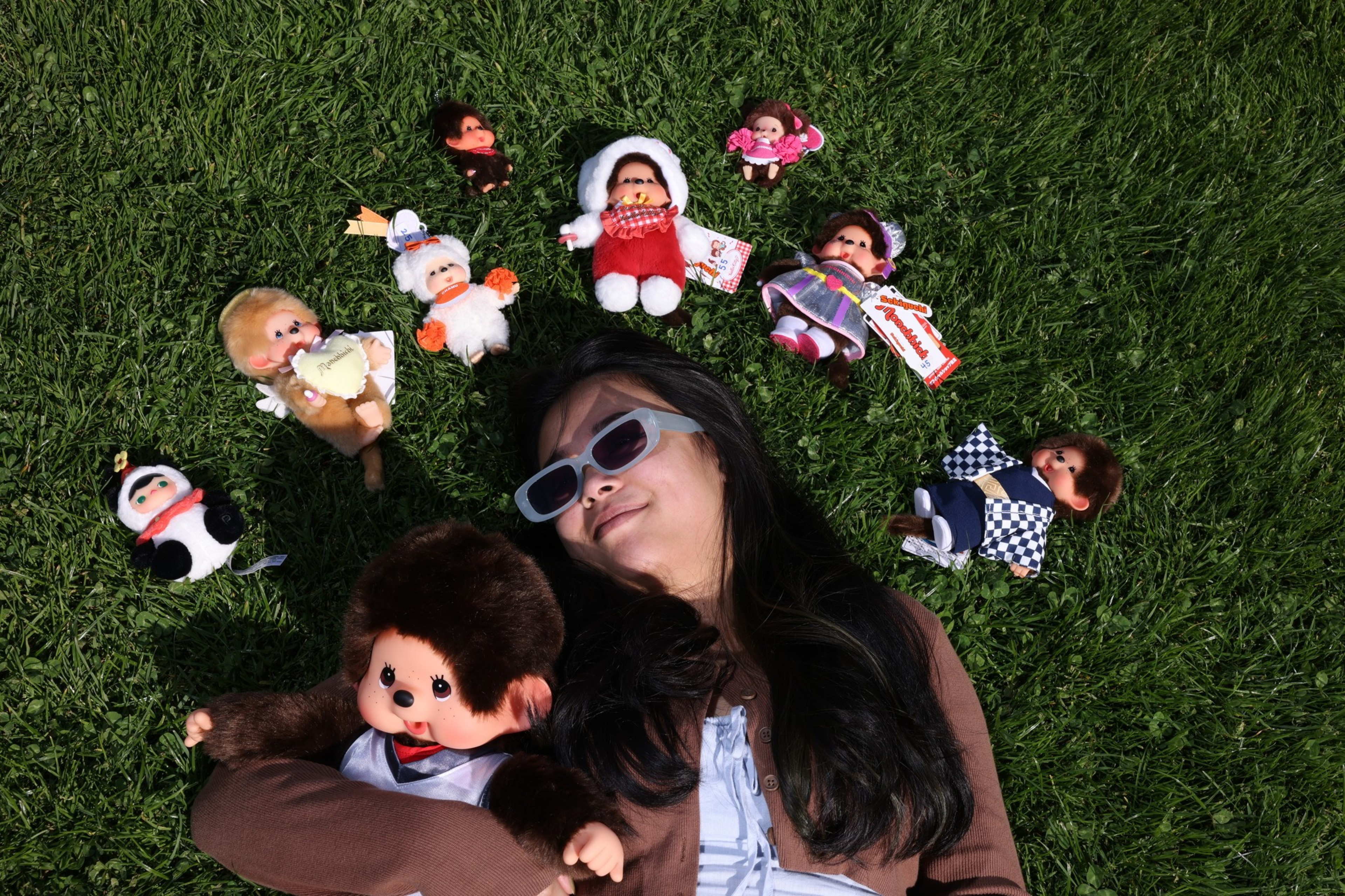 A person wearing sunglasses lies on grass, surrounded by various small plush dolls. The dolls are dressed in different colorful outfits and are arranged in a circle.