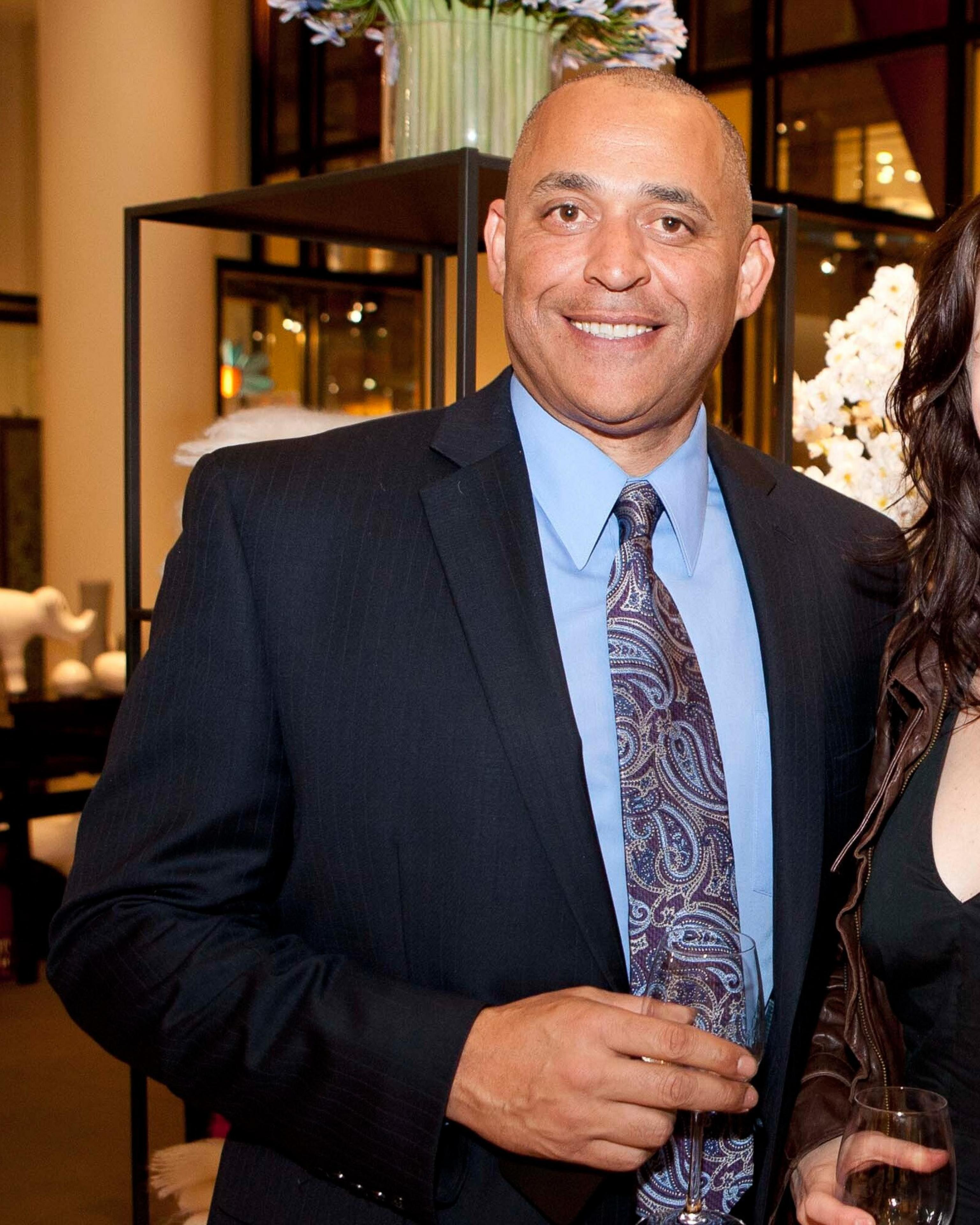 A man in a dark suit with a blue dress shirt and patterned tie is smiling, holding a wine glass, with a floral arrangement and a well-lit, modern background.