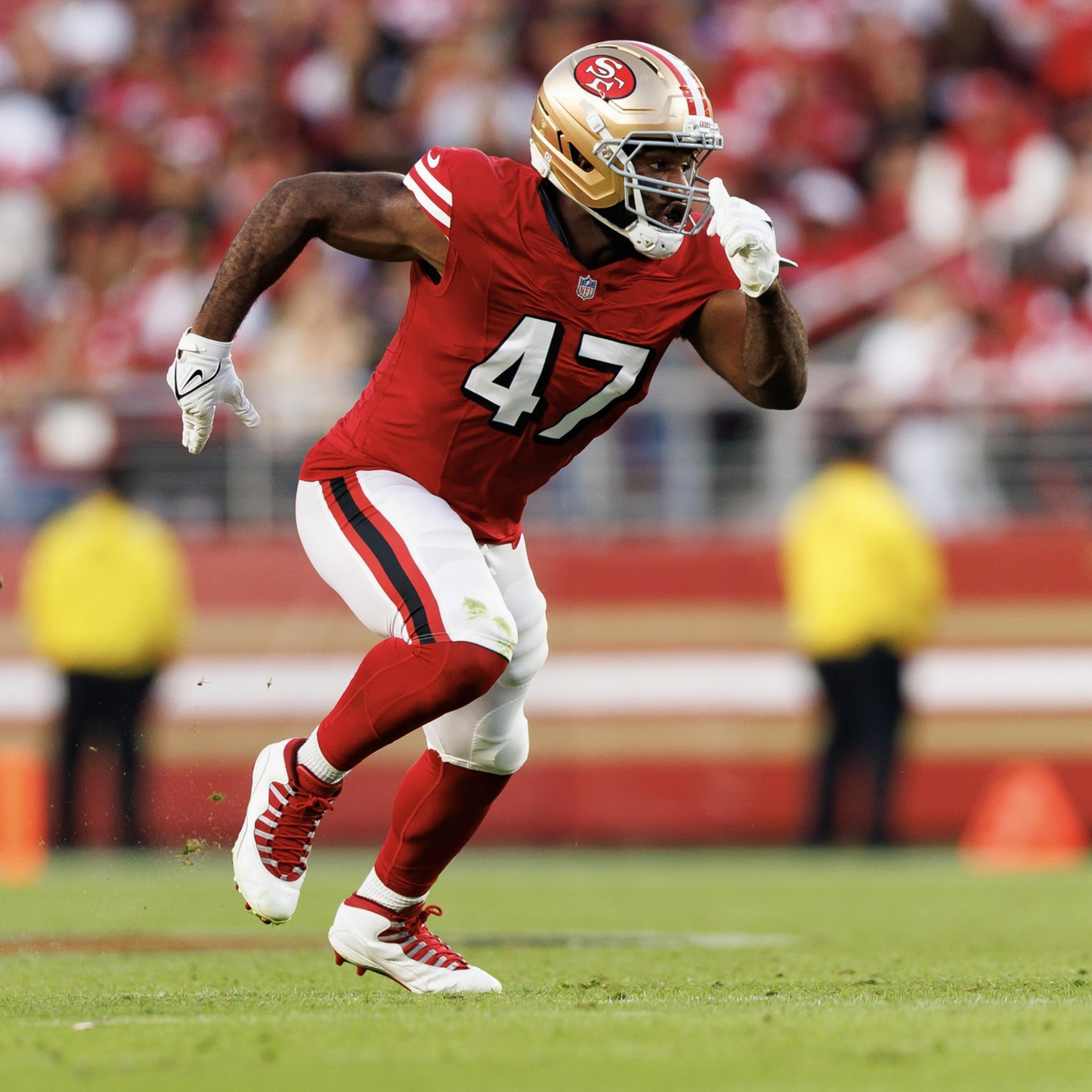 A football player in a red and white uniform with number 47 runs on the field, wearing a gold helmet and white gloves, with a blurred crowd in the background.