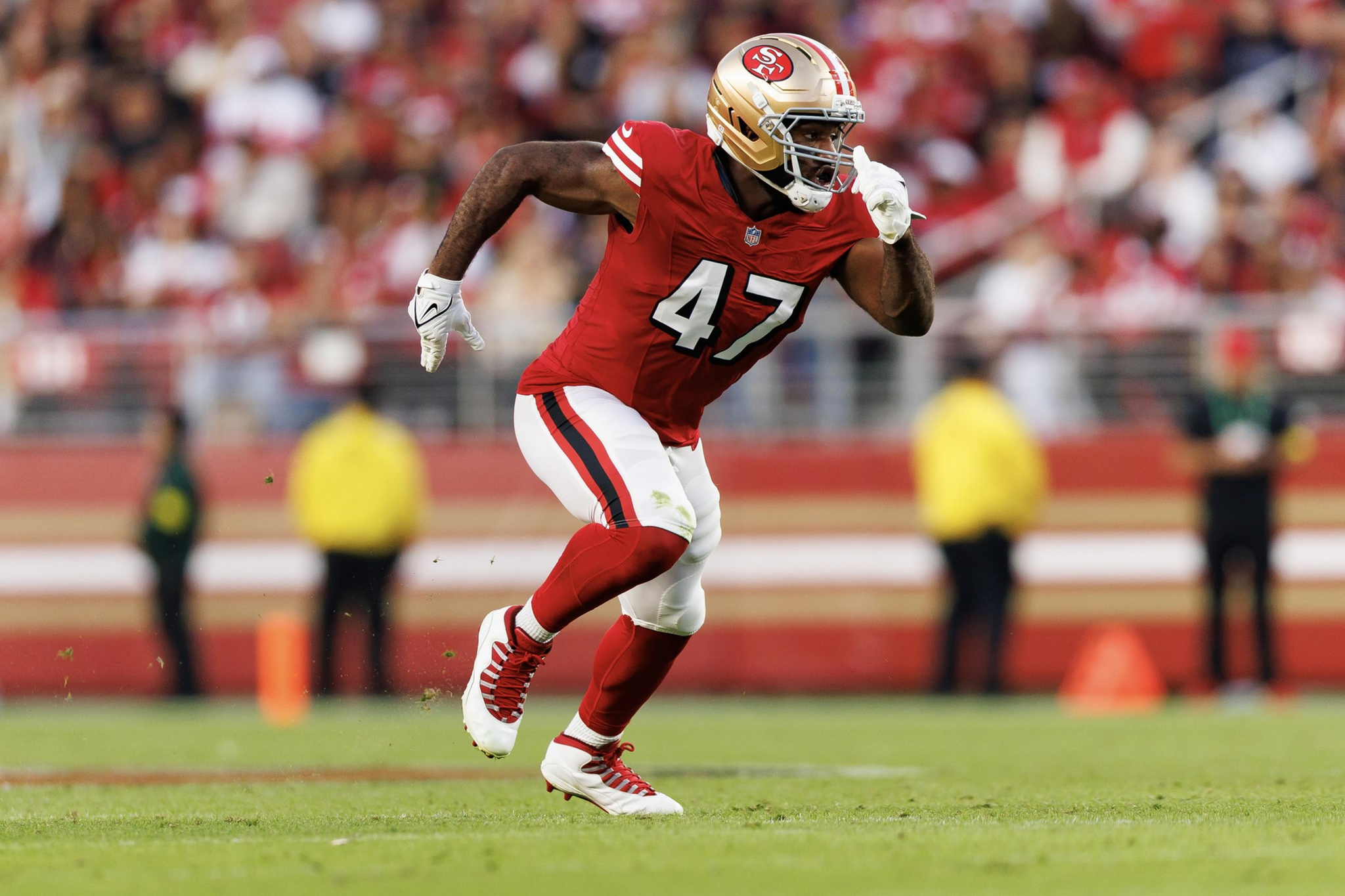 A football player in a red and white uniform with number 47 runs on the field, wearing a gold helmet and white gloves, with a blurred crowd in the background.