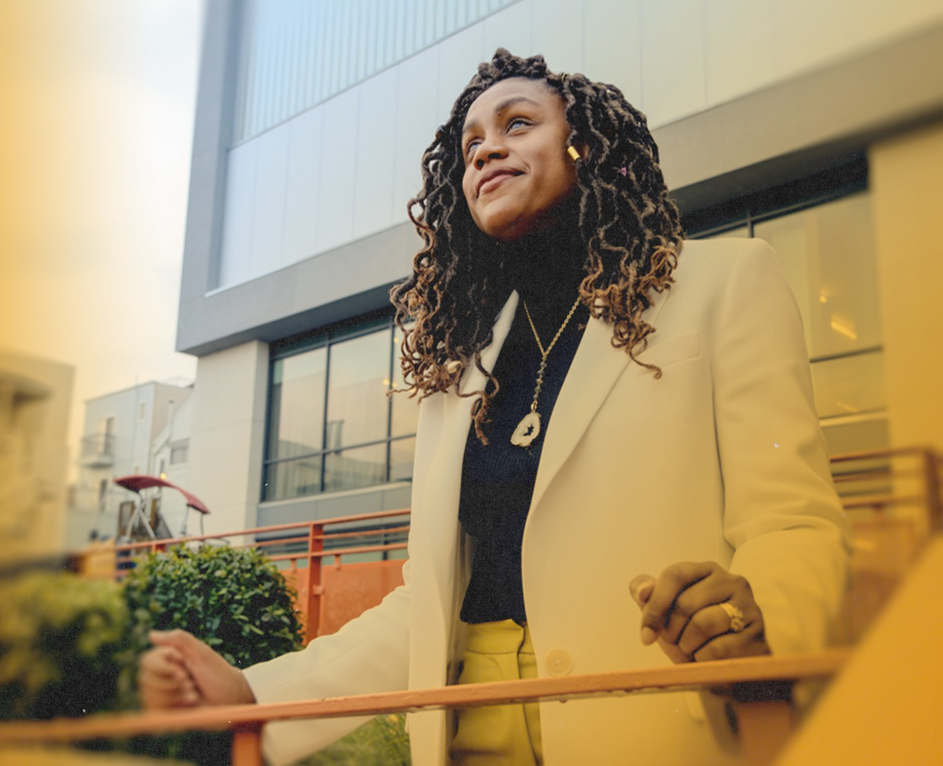 A woman with long curly hair wears a white blazer and black turtleneck, standing outdoors with a calm, confident expression in front of a modern building.