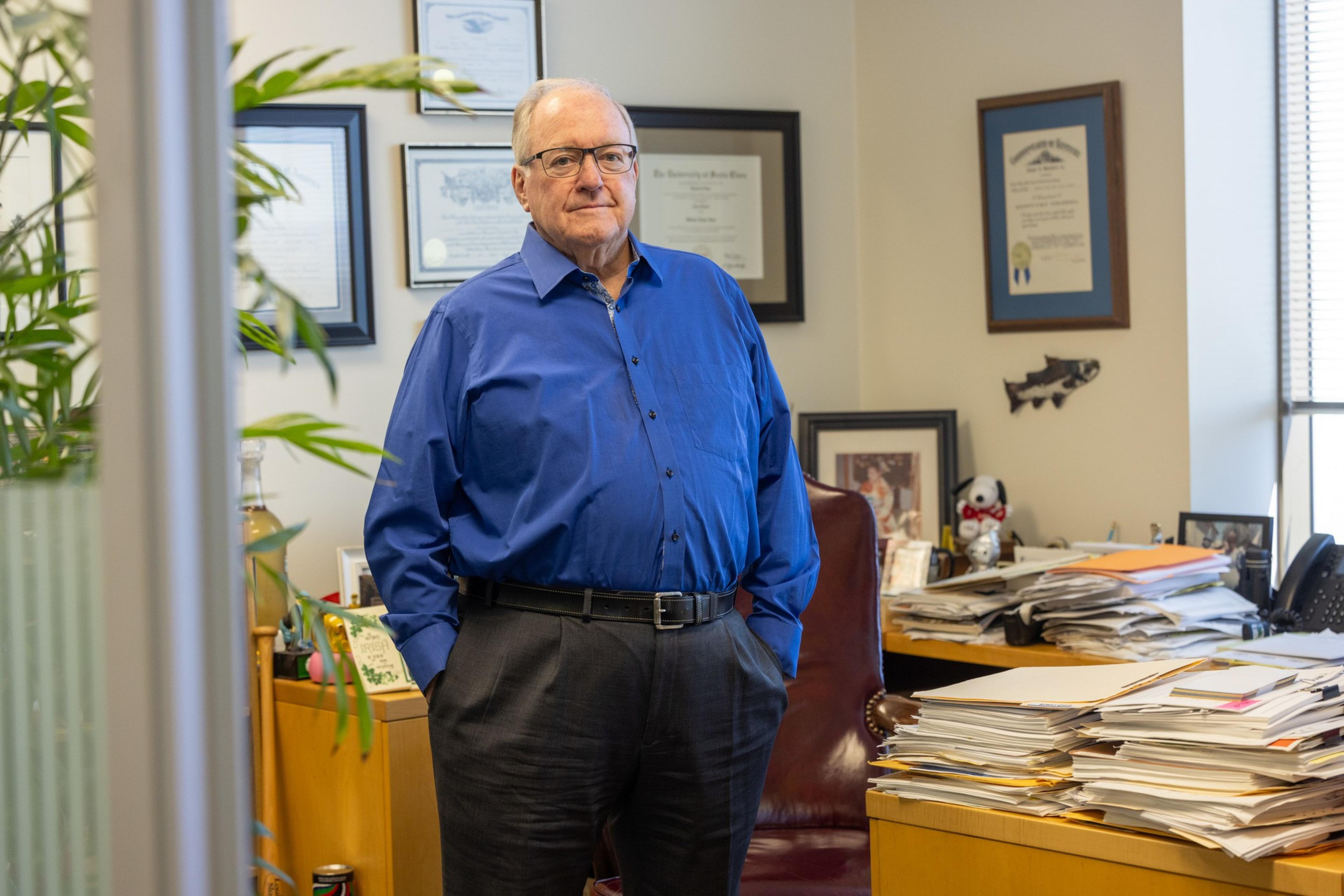 A man in a blue shirt stands in an office filled with framed certificates on the walls, a desk piled with papers, and various decorations and photos.