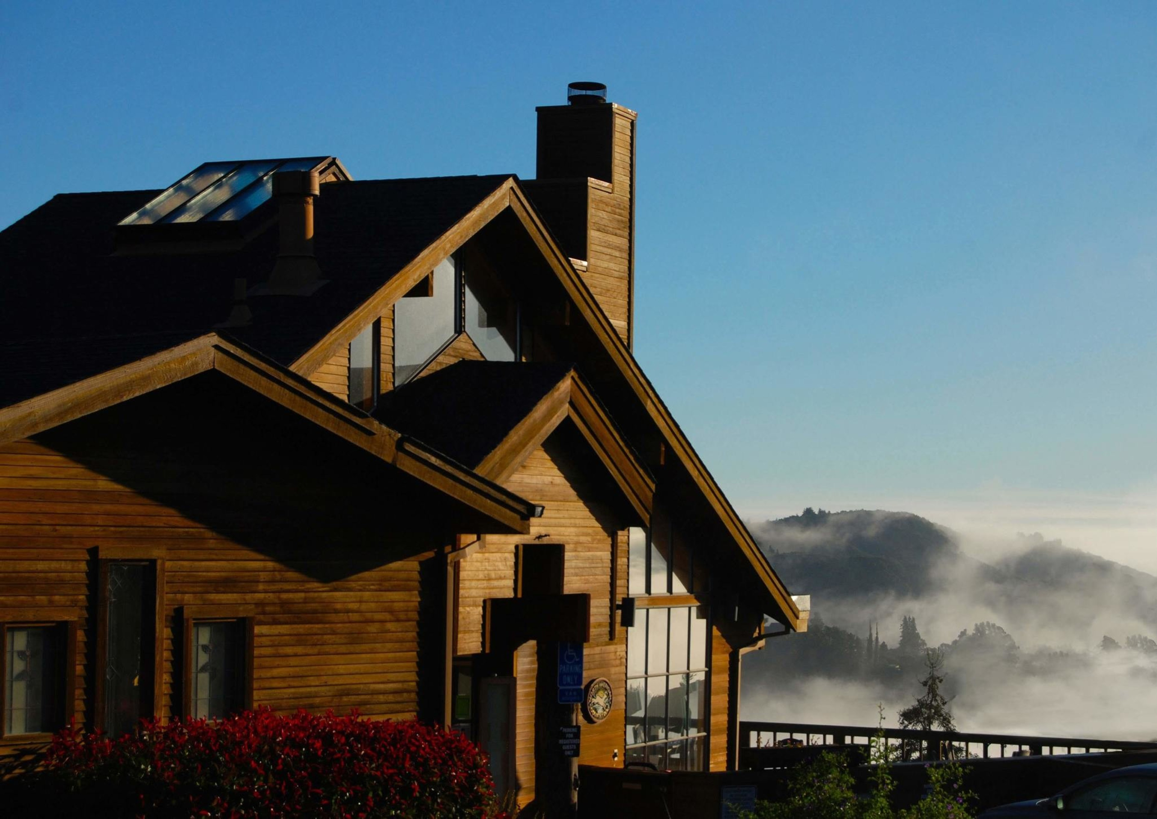 A wooden house with large windows sits on a hillside, overlooking foggy hills under a clear blue sky. A section of a parked car is visible at the bottom.
