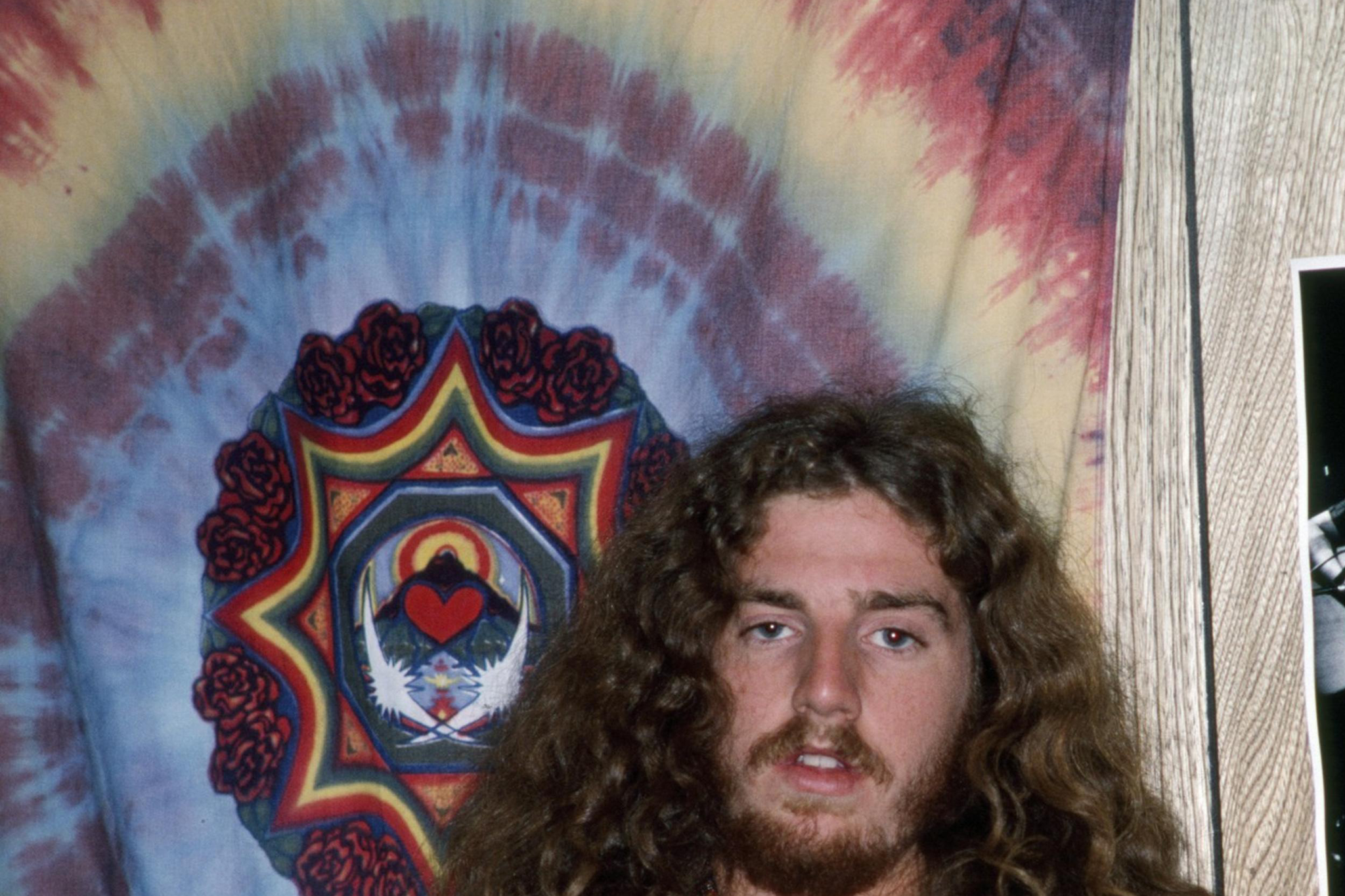 A young man with long curly hair and a beard wears a yellow shirt with embroidered flowers, sitting in front of a tie-dye tapestry and a black-and-white musician photo.