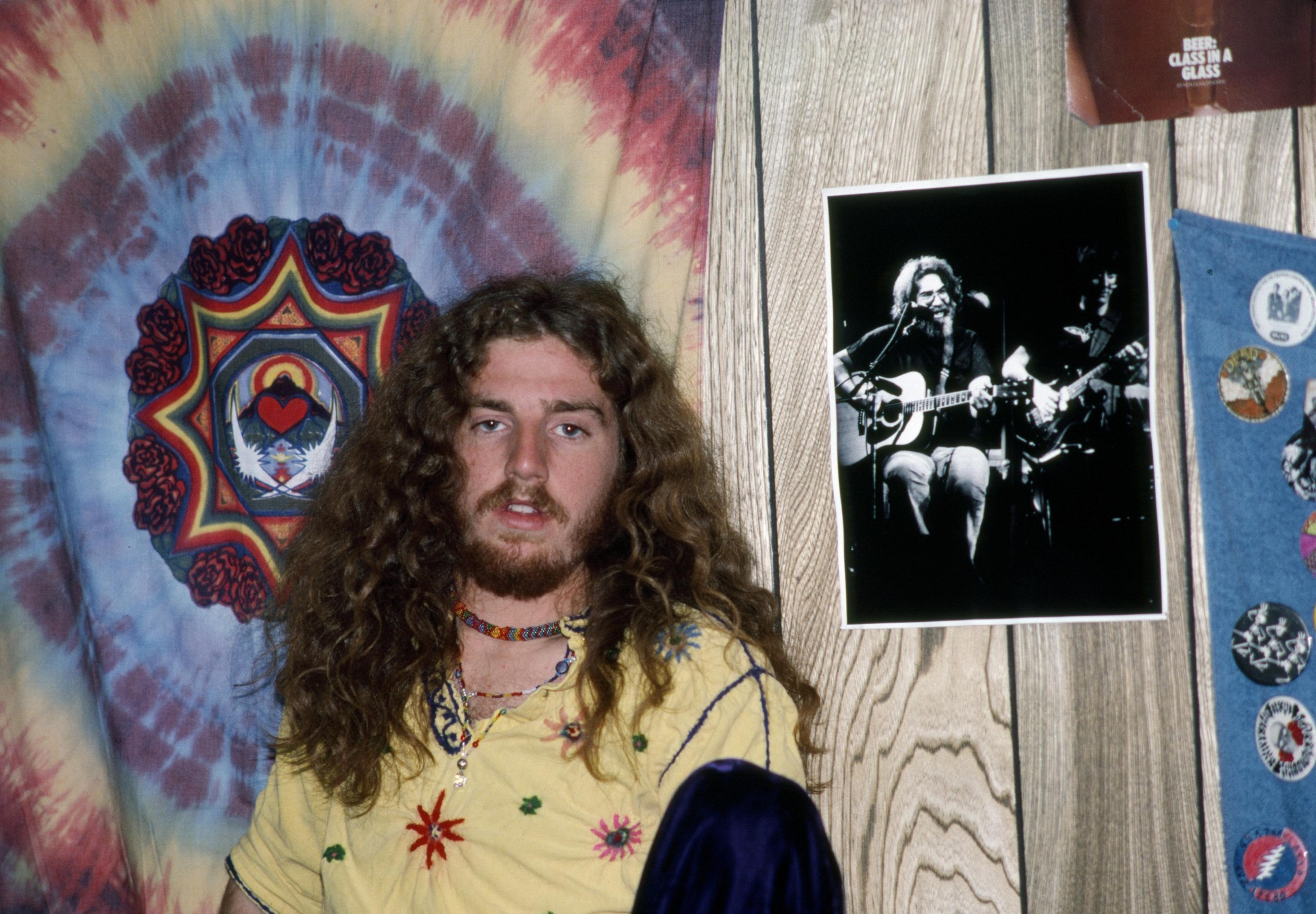 A young man with long curly hair and a beard wears a yellow shirt with embroidered flowers, sitting in front of a tie-dye tapestry and a black-and-white musician photo.