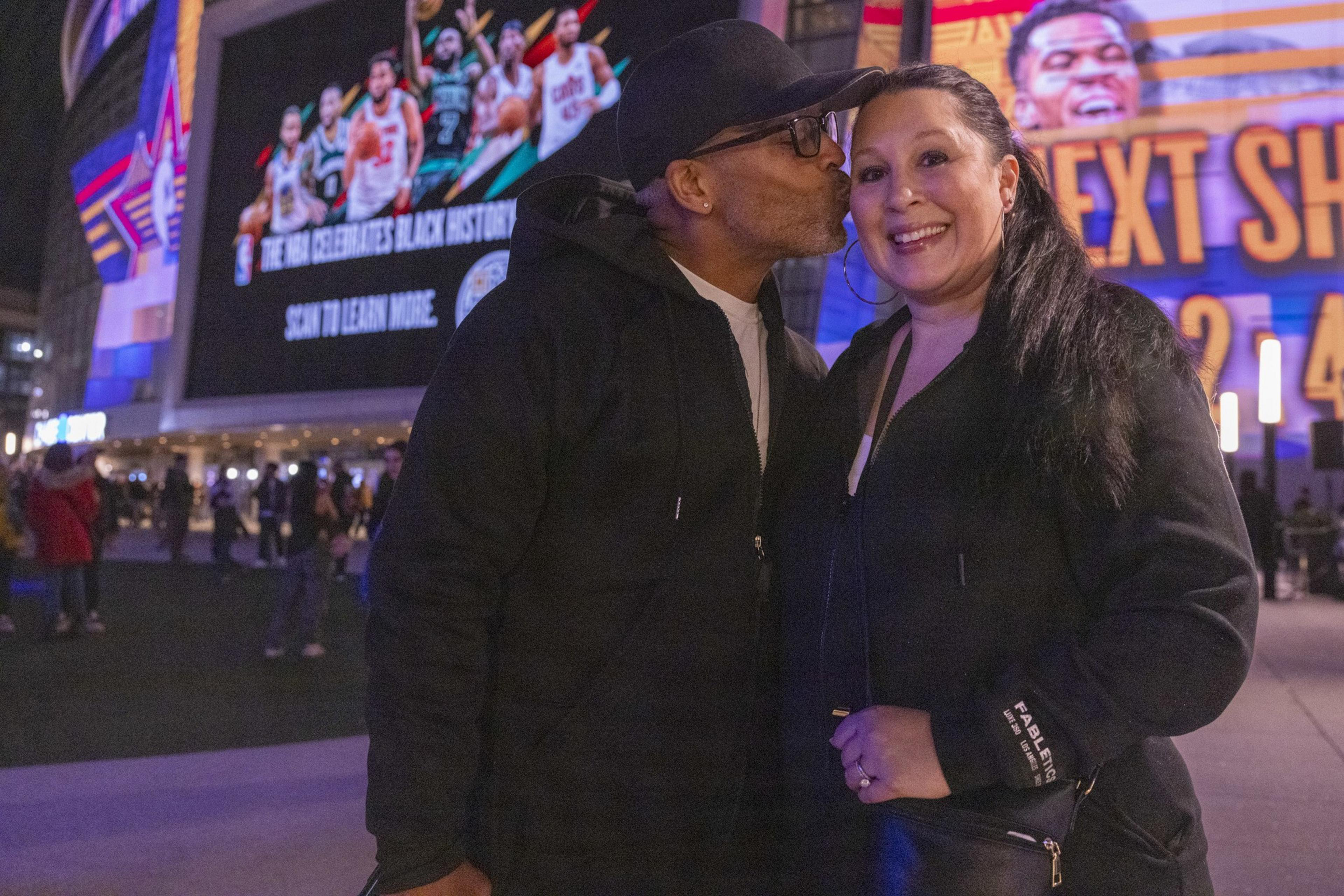 A man in a cap kisses a smiling woman on the cheek. They're outdoors near a large, colorful billboard displaying basketball players and text.