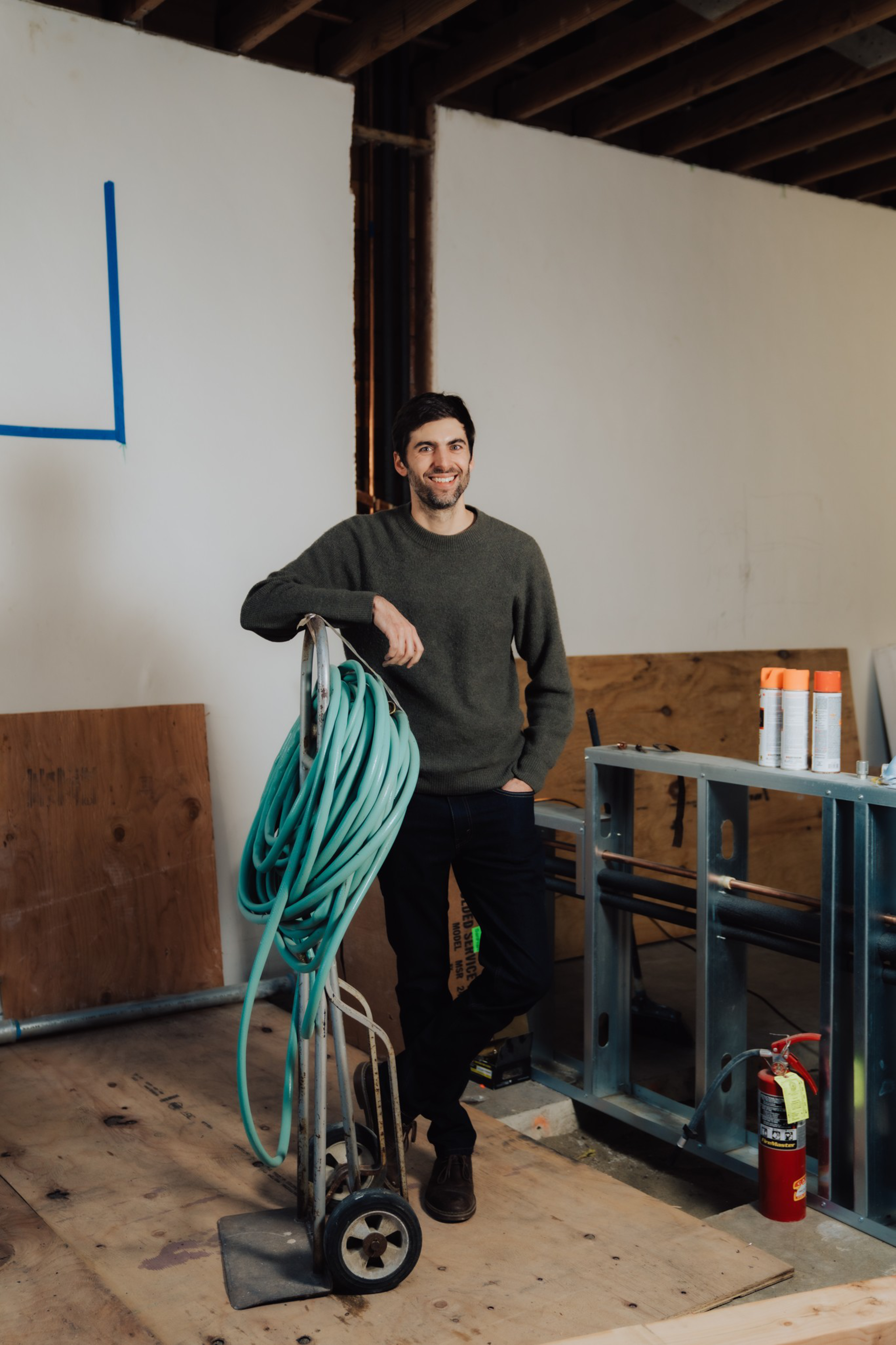 A smiling man in a dark sweater and pants leans on a cart with coiled green hose in an unfinished room with plywood and a fire extinguisher.