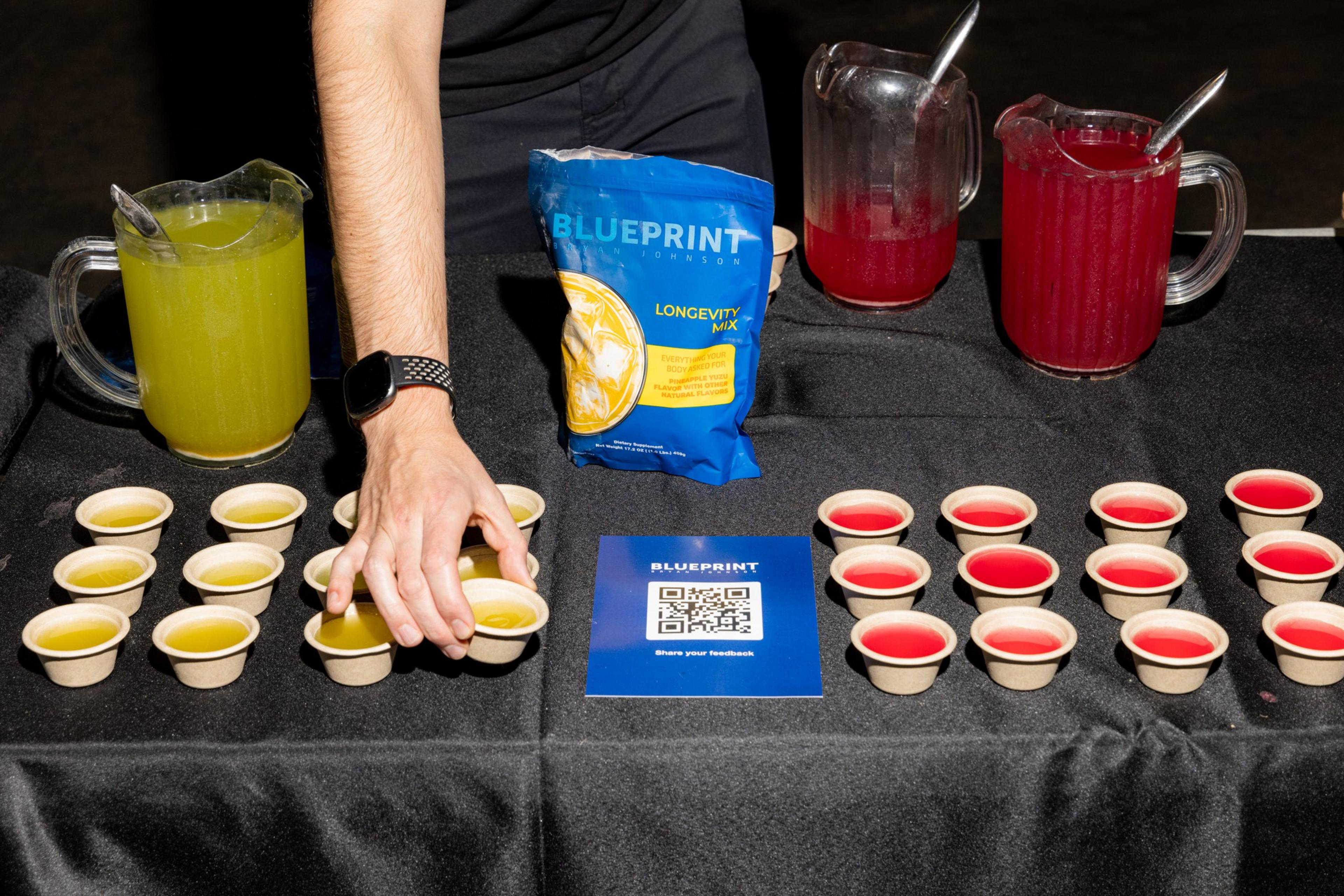 A person is placing small cups of yellow and red drinks on a table with pitchers of the same drinks behind them. A package labeled &quot;BLUEPRINT&quot; and a QR code are also visible.