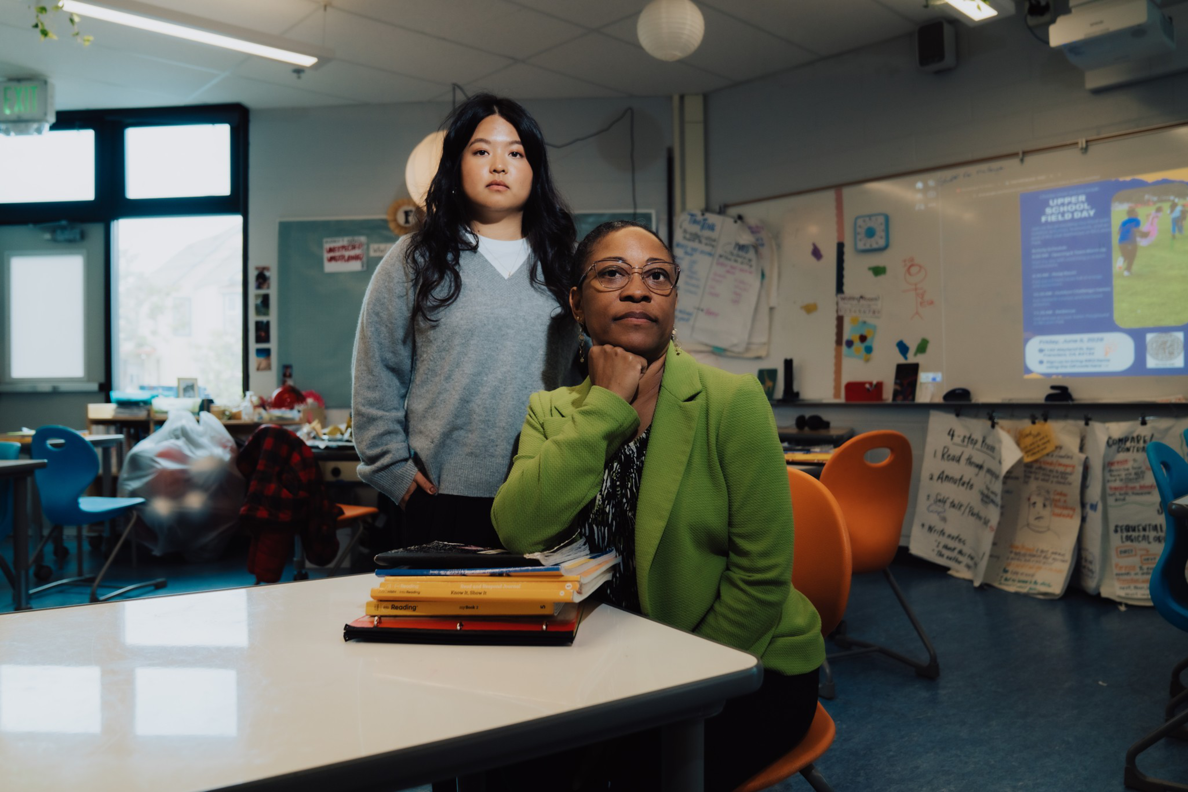 One woman in a green blazer sits at a desk with books, resting her chin on her hand, while another woman in a gray sweater stands behind her in a classroom.