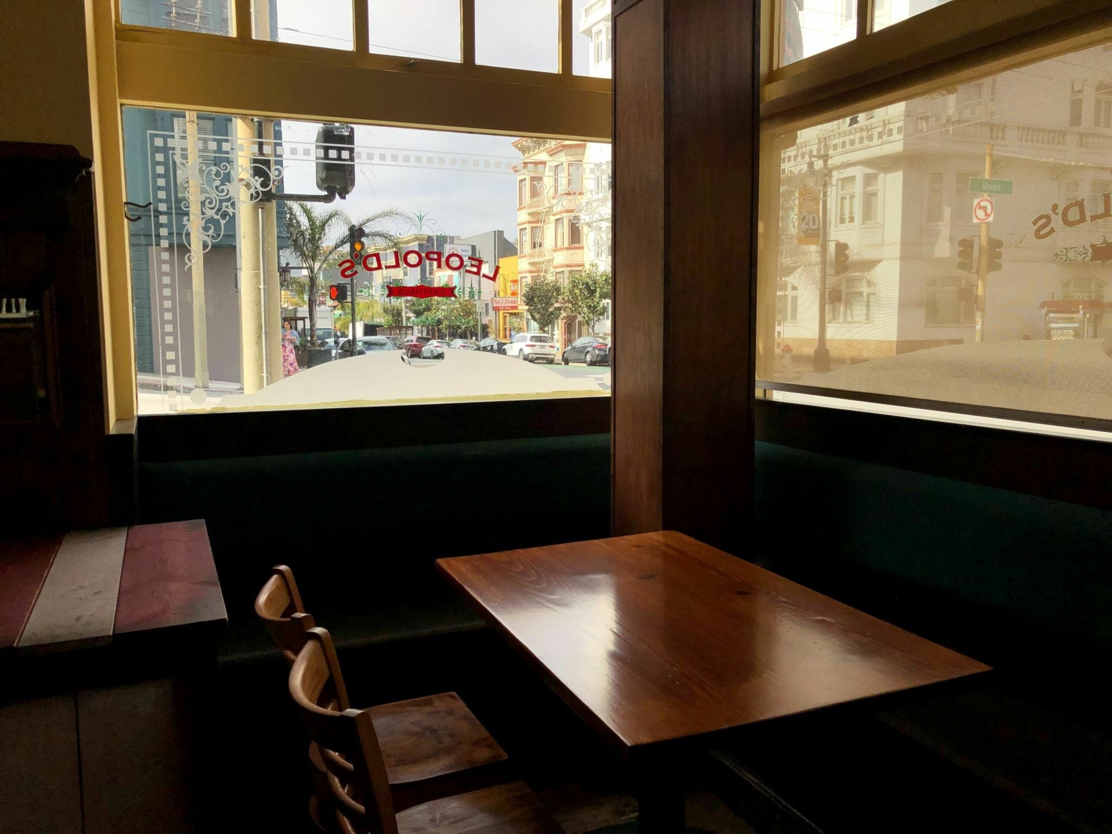 A wooden table with two chairs and a bench sits by a large window with the word "Leopold's" in red and a view of a sunny street with buildings and palm trees.