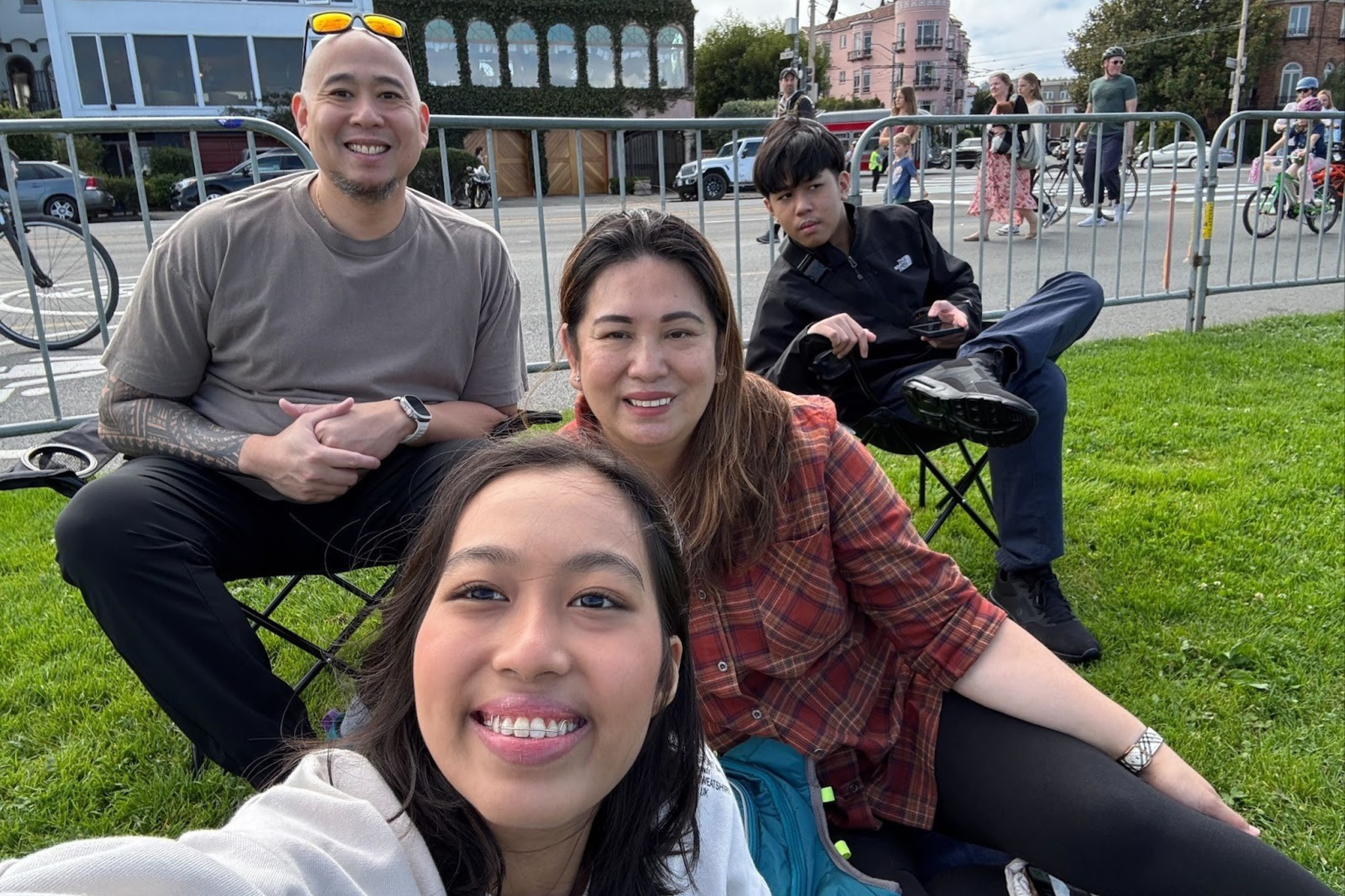 A group of four people, two men and two women, sit on a grassy area by a street. They look relaxed and happy, with a few pedestrians in the background.