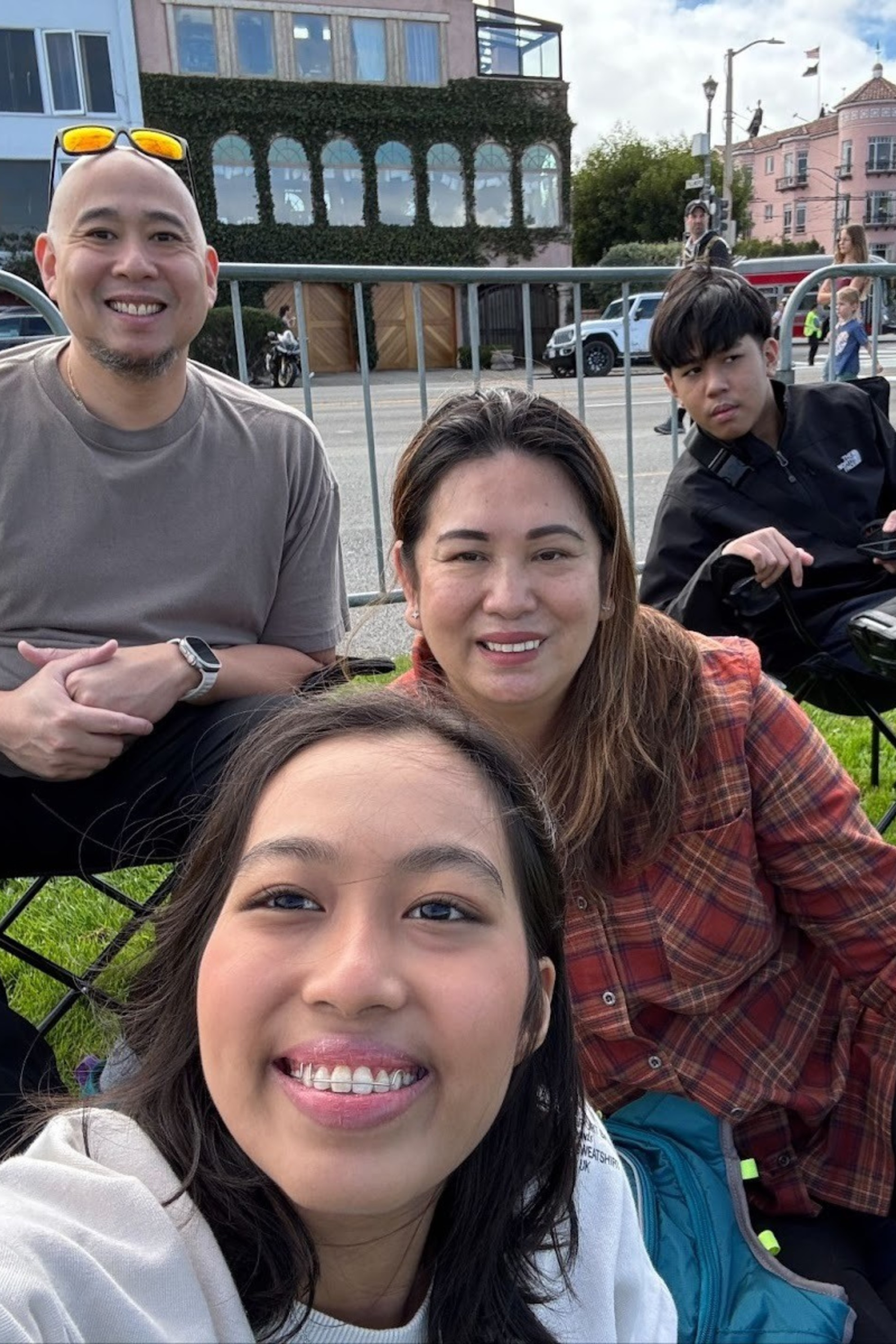 A group of four people, two men and two women, sit on a grassy area by a street. They look relaxed and happy, with a few pedestrians in the background.