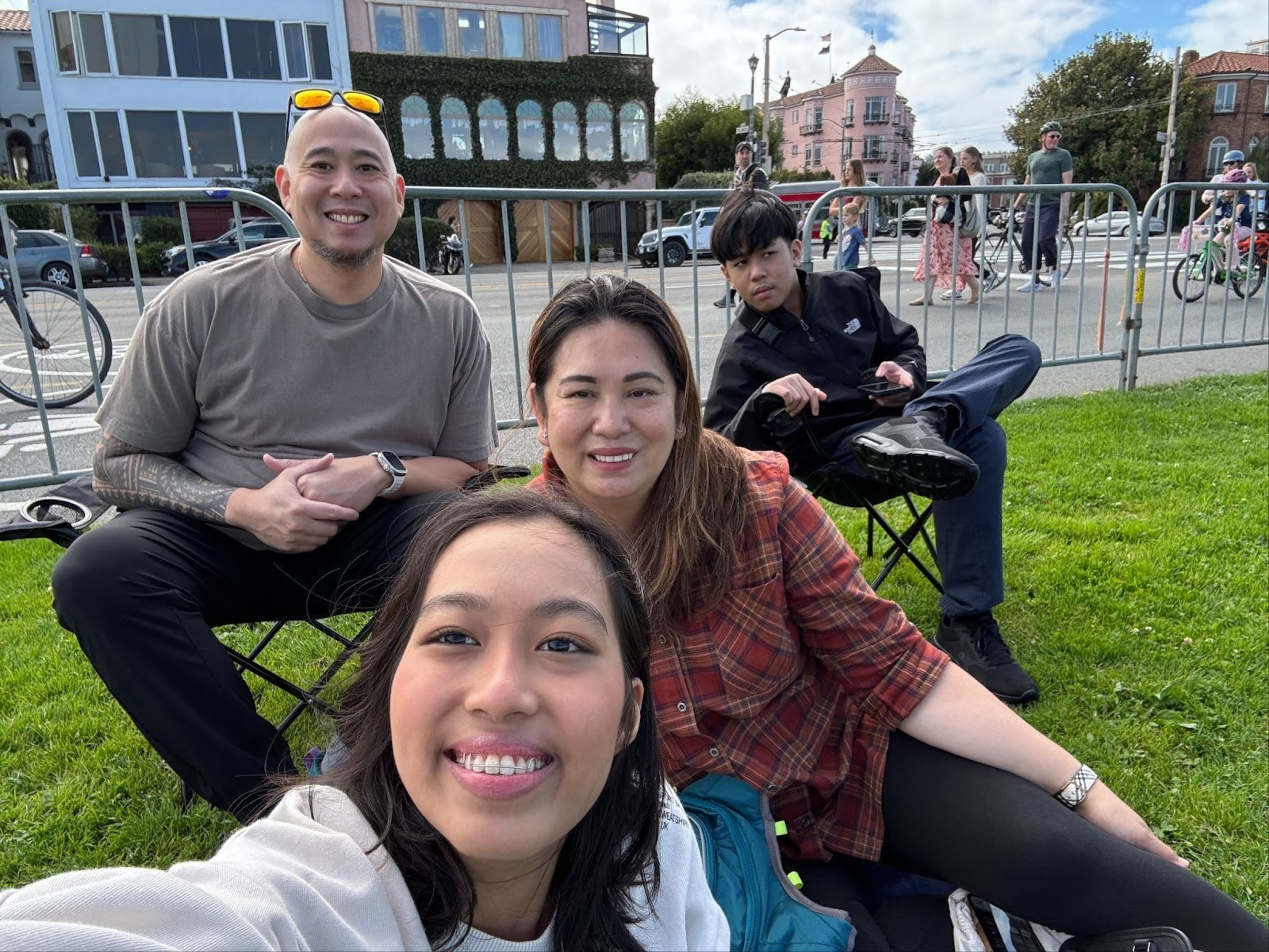 A group of four people, two men and two women, sit on a grassy area by a street. They look relaxed and happy, with a few pedestrians in the background.