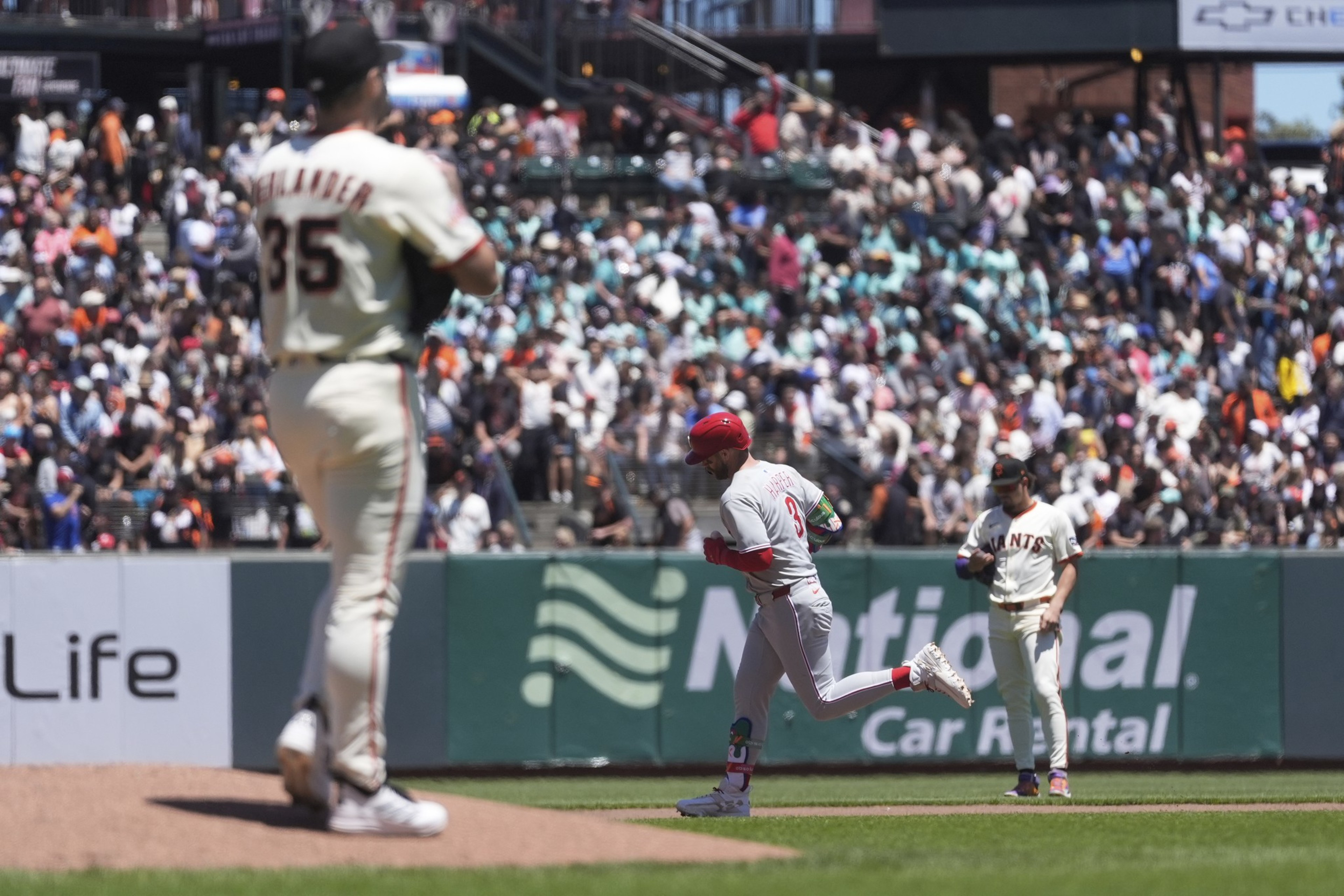 A baseball game is captured with a player in a white uniform standing on the pitcher's mound, another in a gray uniform running past, and a crowd in the background.