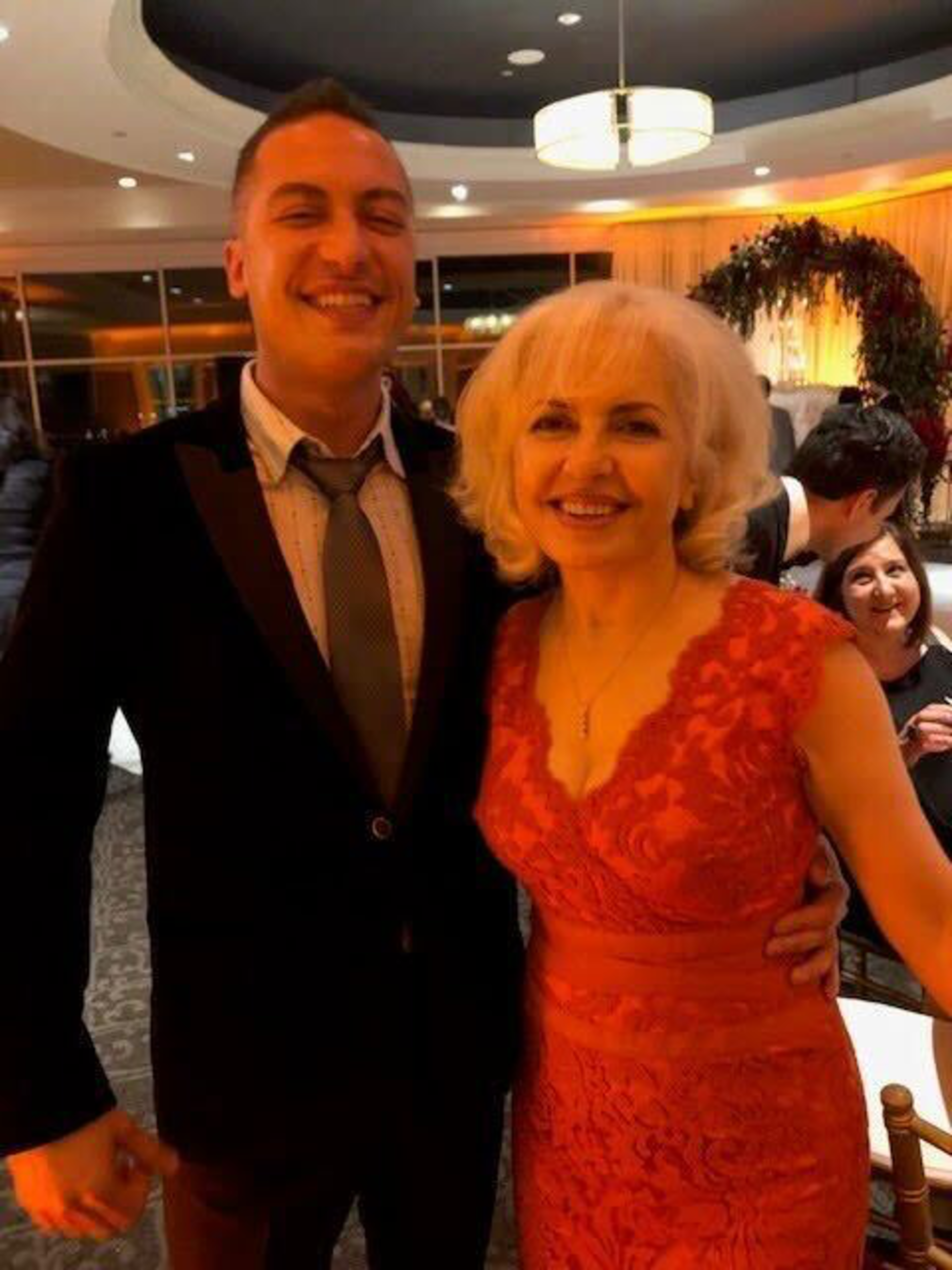 A smiling man in a dark suit and gray tie stands with a smiling woman in a red lace dress at a warmly lit indoor event.