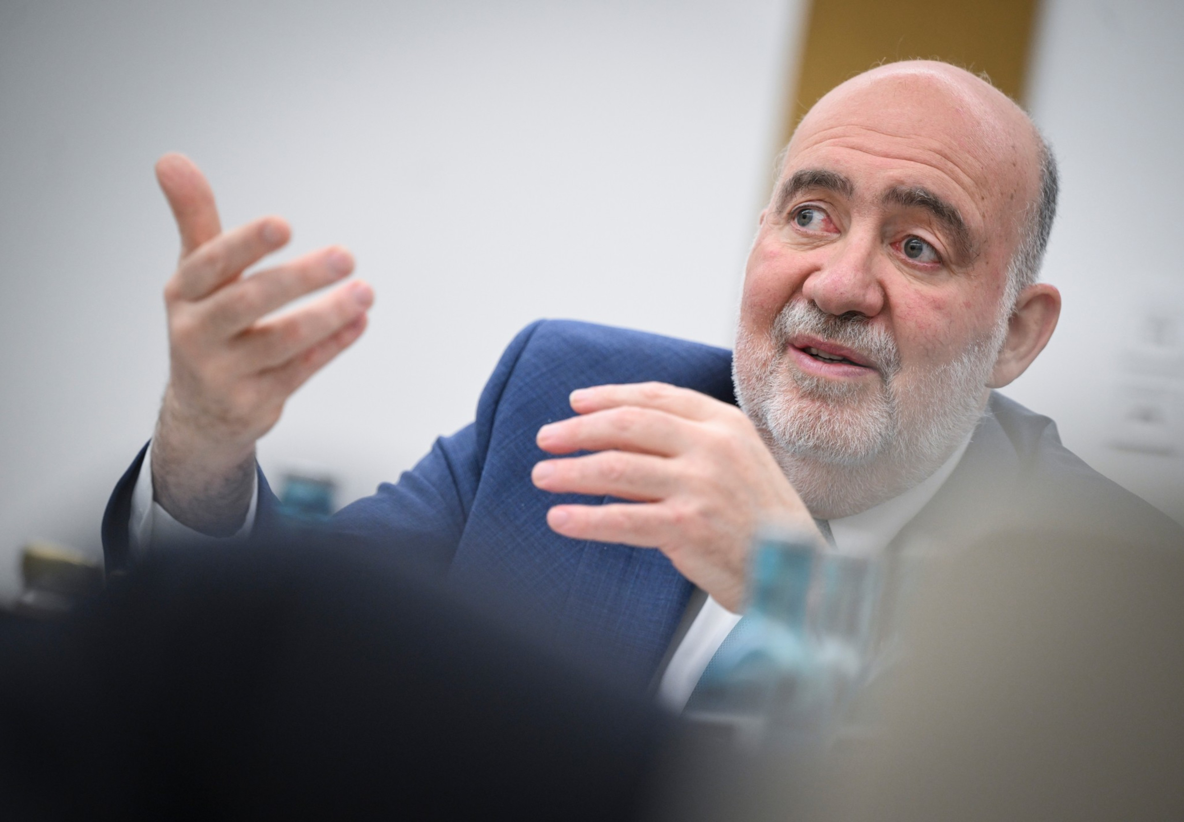 A bearded man in a blue suit gestures with both hands while speaking, with a focused and engaged expression.