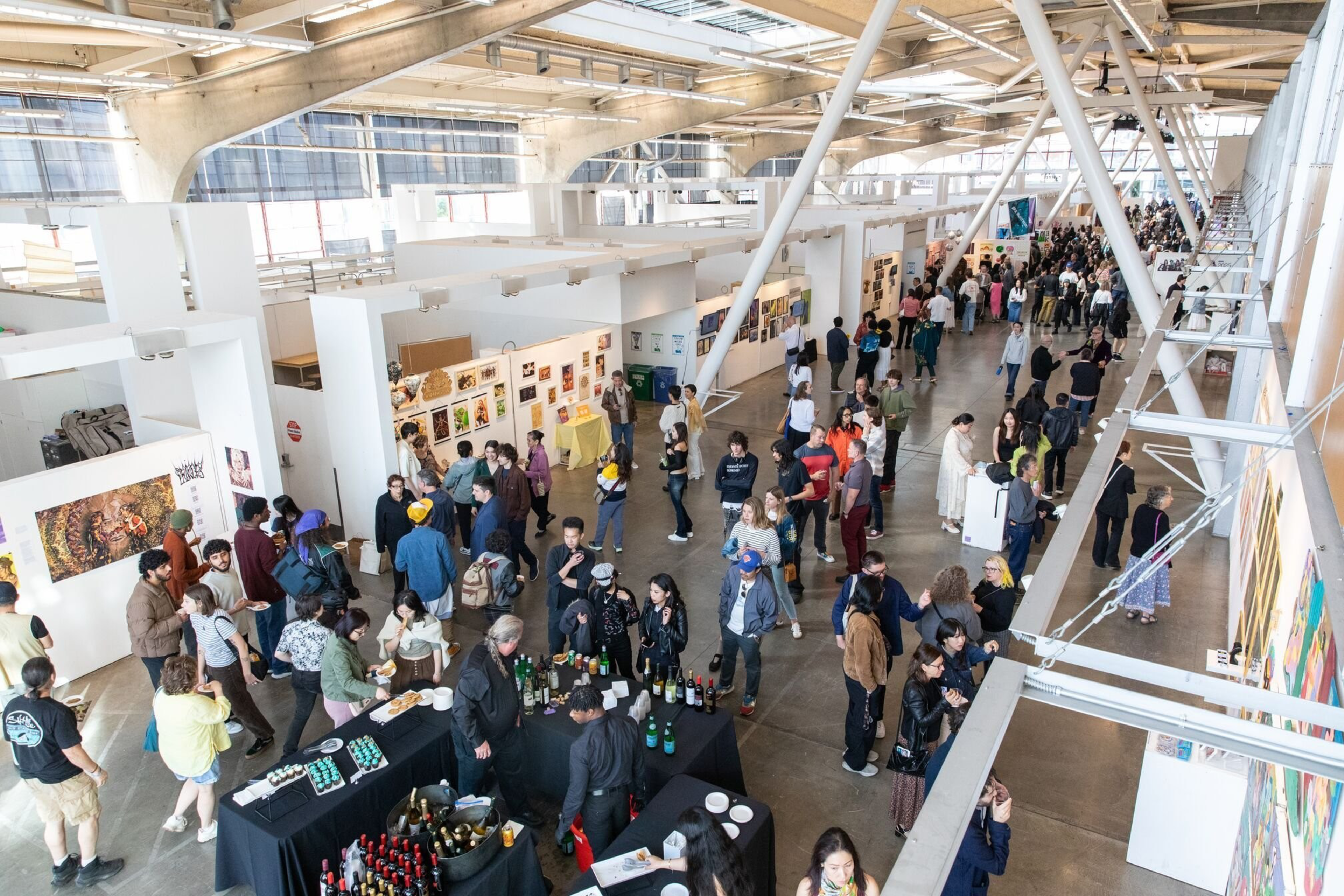 A spacious, industrial-style gallery hosts an art exhibit with many visitors viewing colorful artwork on white walls and mingling near a refreshment table.