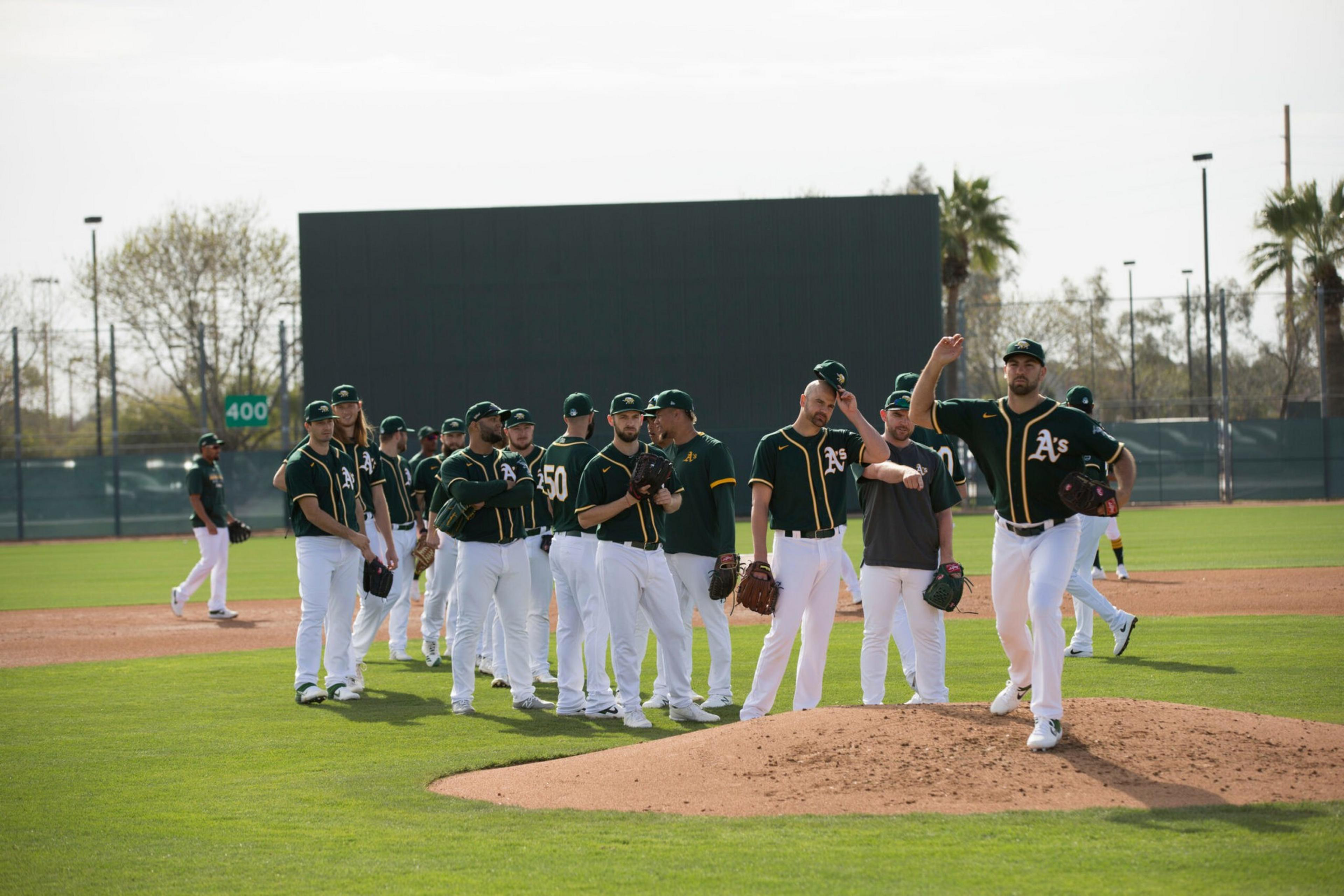 A group of baseball players in green and yellow uniforms stands on the field, with one pitcher mid-throw on the mound.