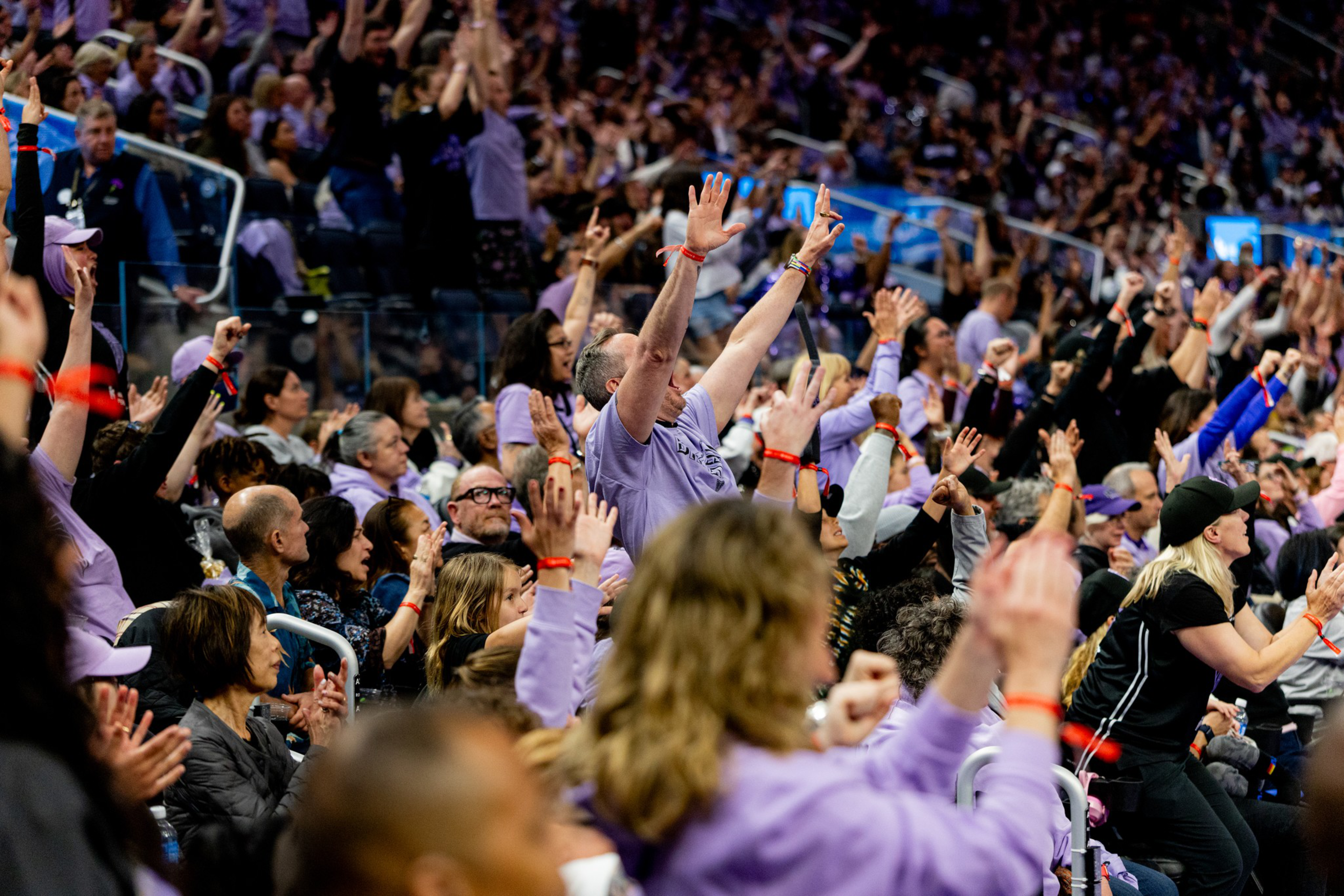A large crowd of people, many wearing purple, are raising their hands and cheering enthusiastically in a stadium or arena.
