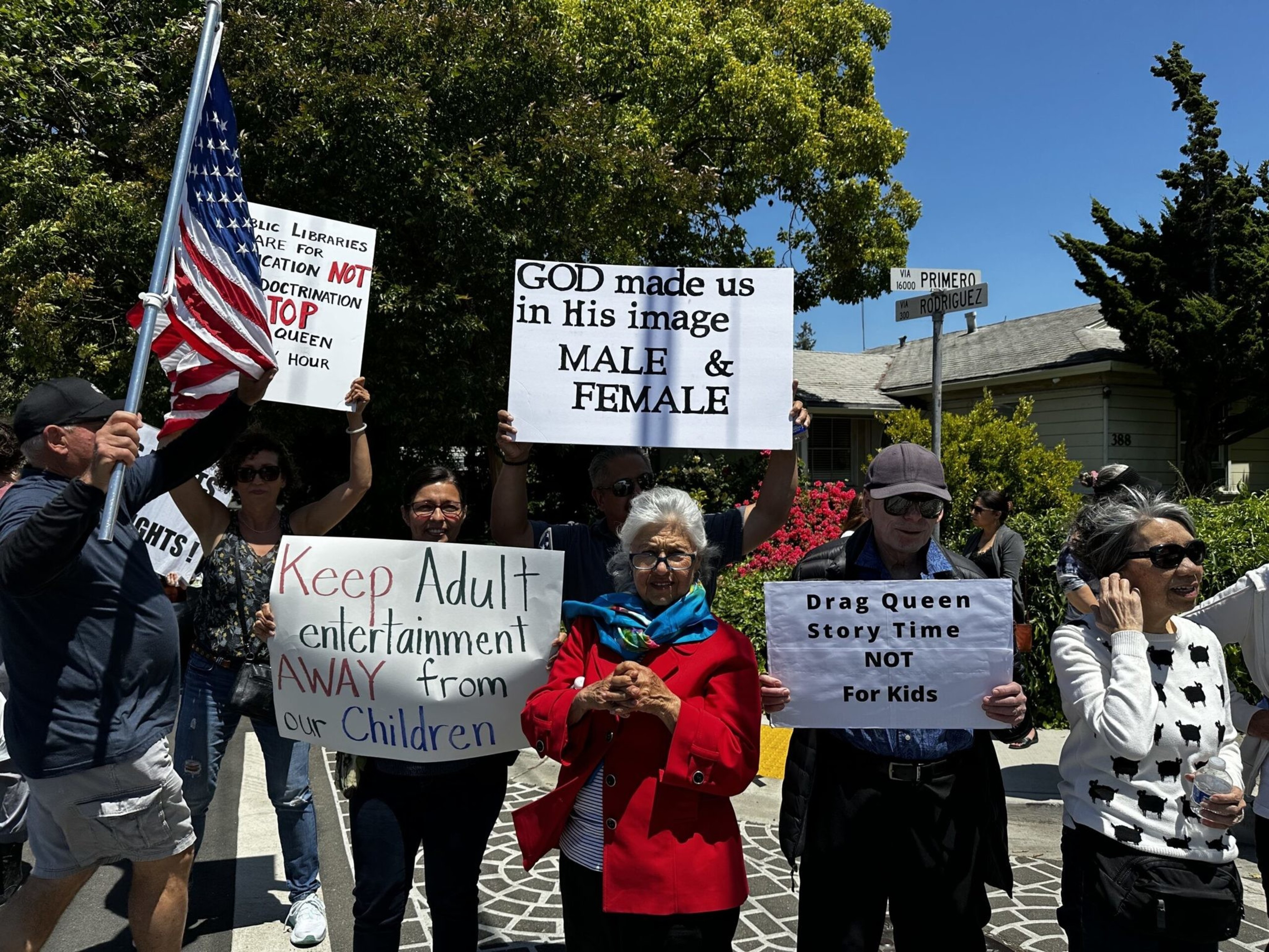 A group of people hold signs opposing drag queen story time in libraries, with messages about gender, children, and education, while one holds an American flag.