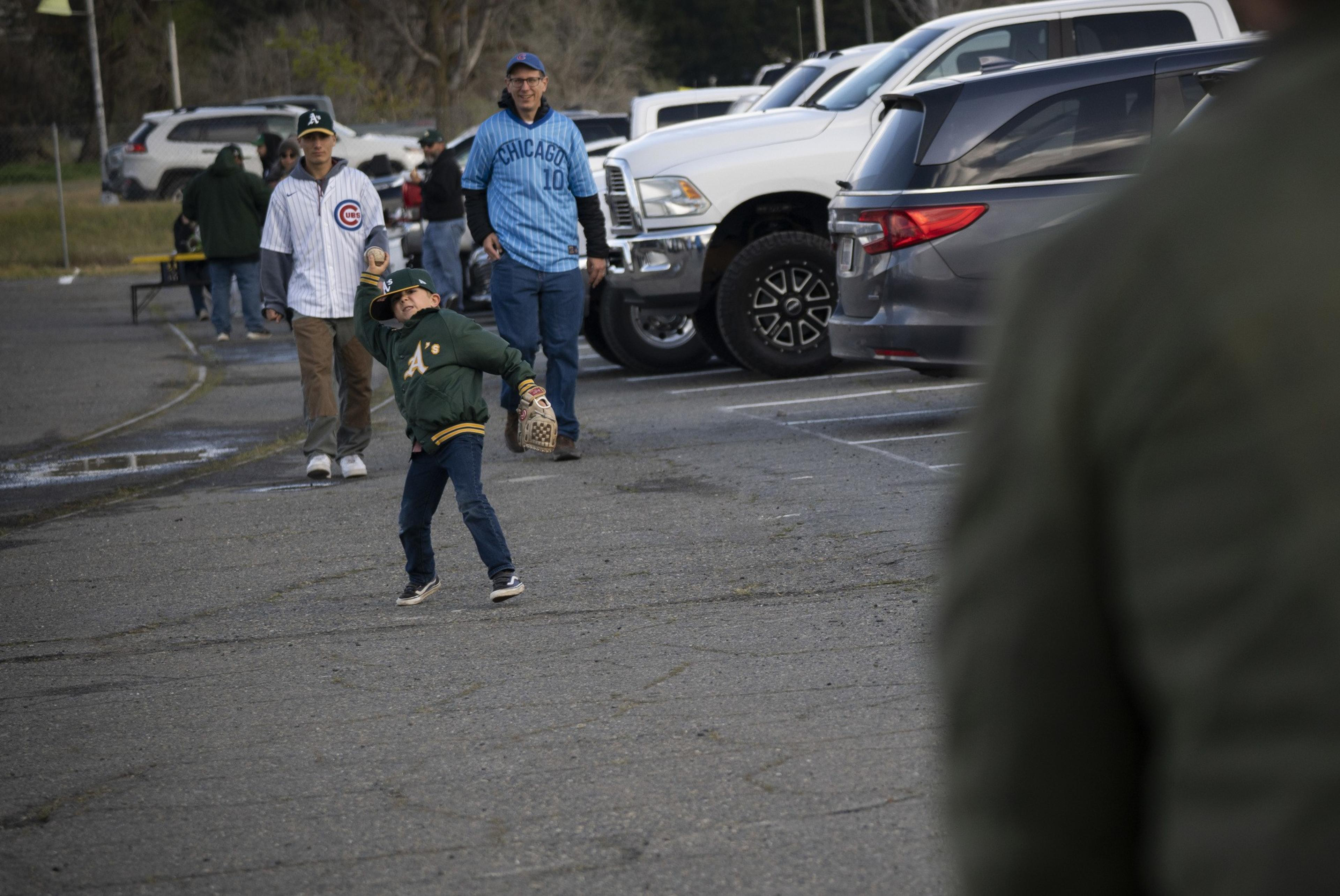 A child in an A's jacket throws a baseball in a parking lot, while two adults in sports jerseys watch near parked cars.