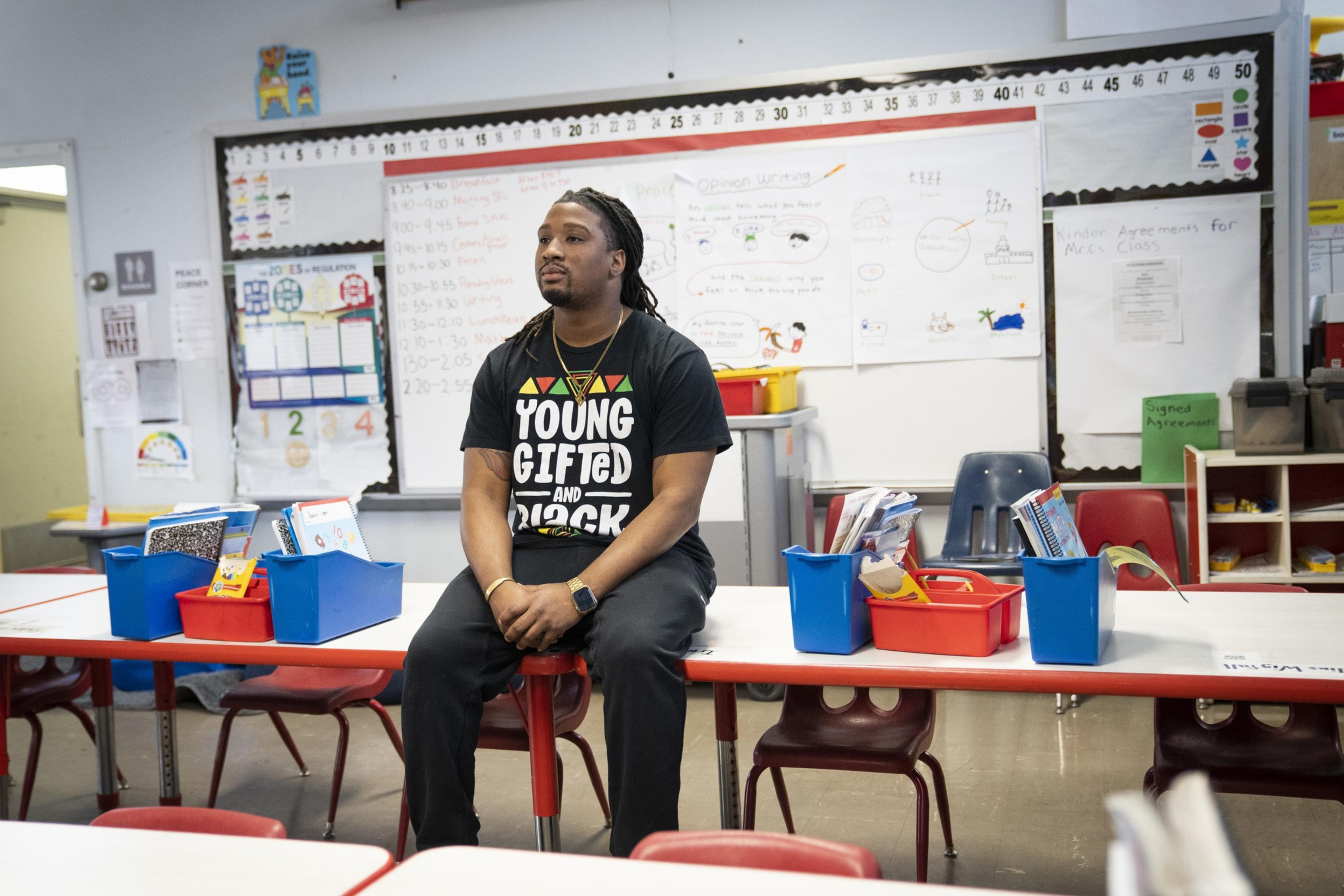 A man wearing a "Young, Gifted and Black" shirt sits on a school desk in a classroom with colorful bins and educational posters around him.