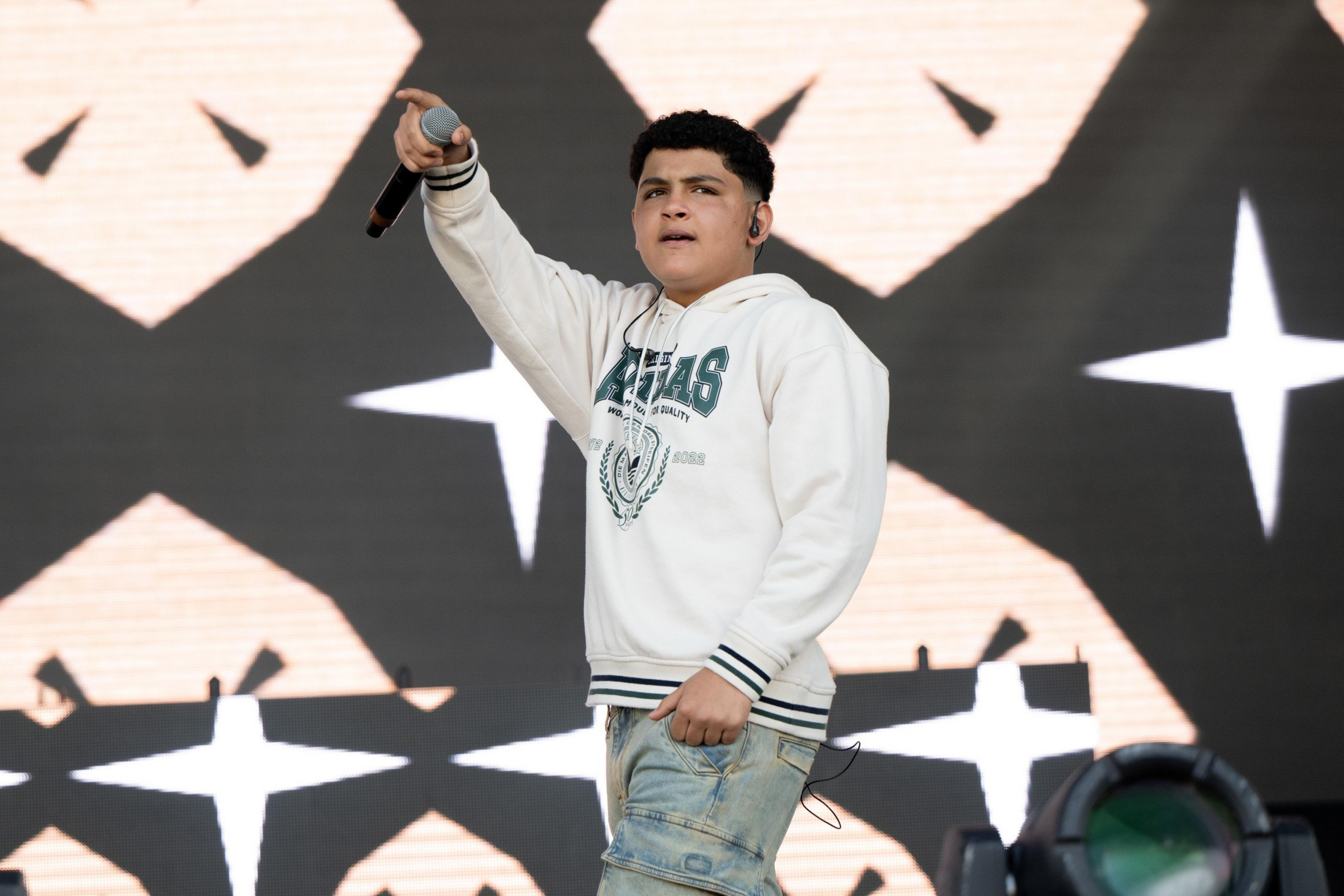 A person performing on stage holds a microphone, wearing a white hoodie and light jeans. The background features a geometric pattern with star-like designs.