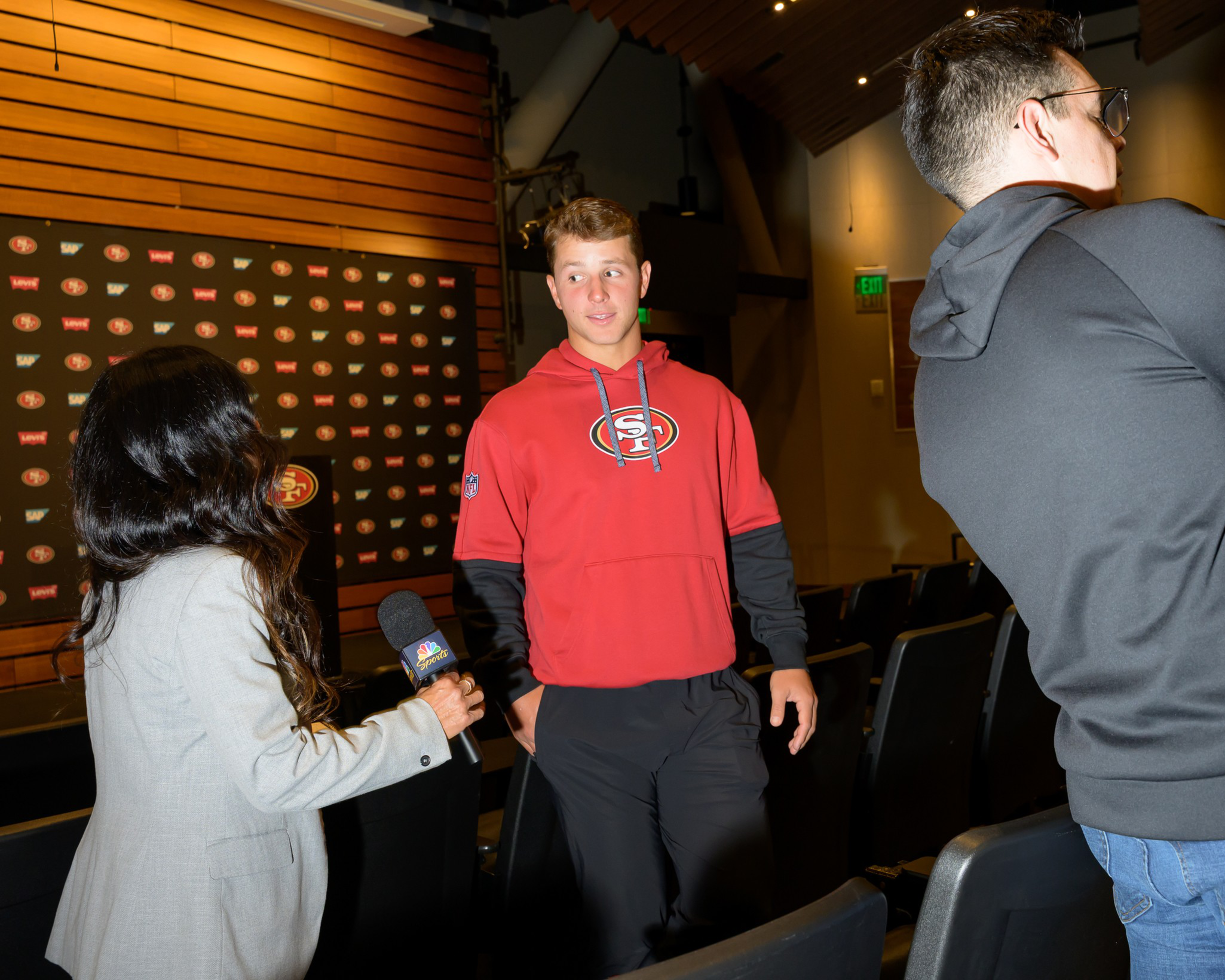 A young man in a red sports hoodie stands in a room facing a woman holding a microphone. A man in a black hoodie is partially visible on the right.