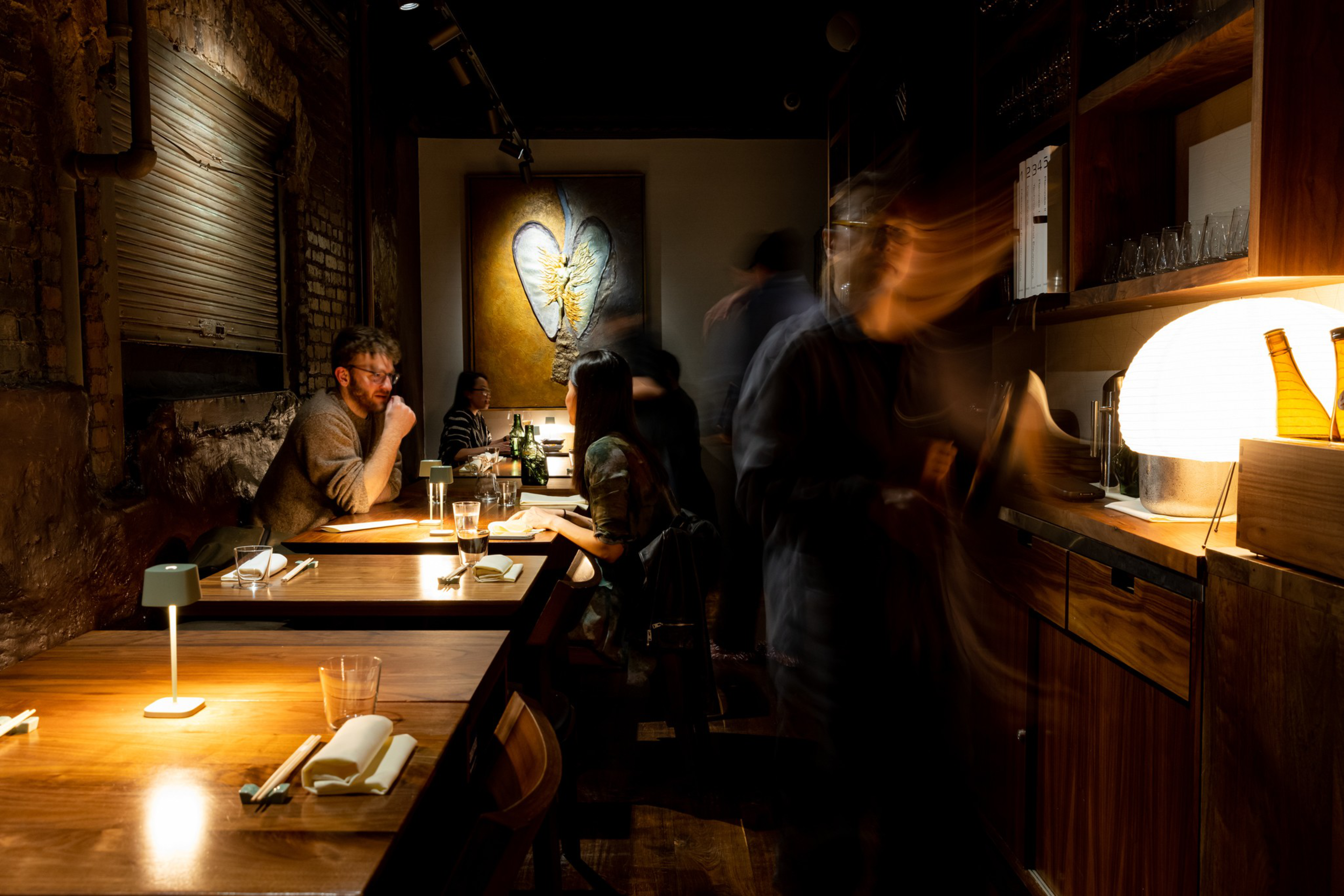 A dimly lit restaurant with wooden tables hosts two seated couples, while blurred figures move near a glowing lamp on a wooden counter.