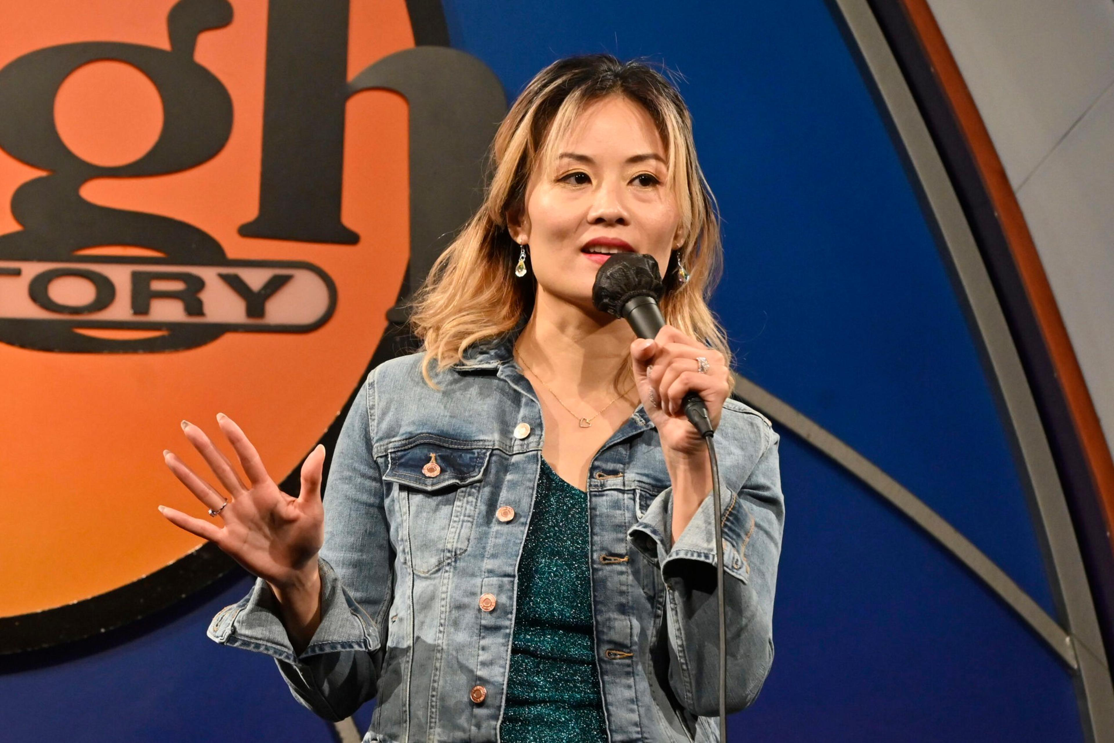A woman wearing a denim jacket and sparkly top holds a microphone and gestures with her hand on stage in front of a large orange and black sign.