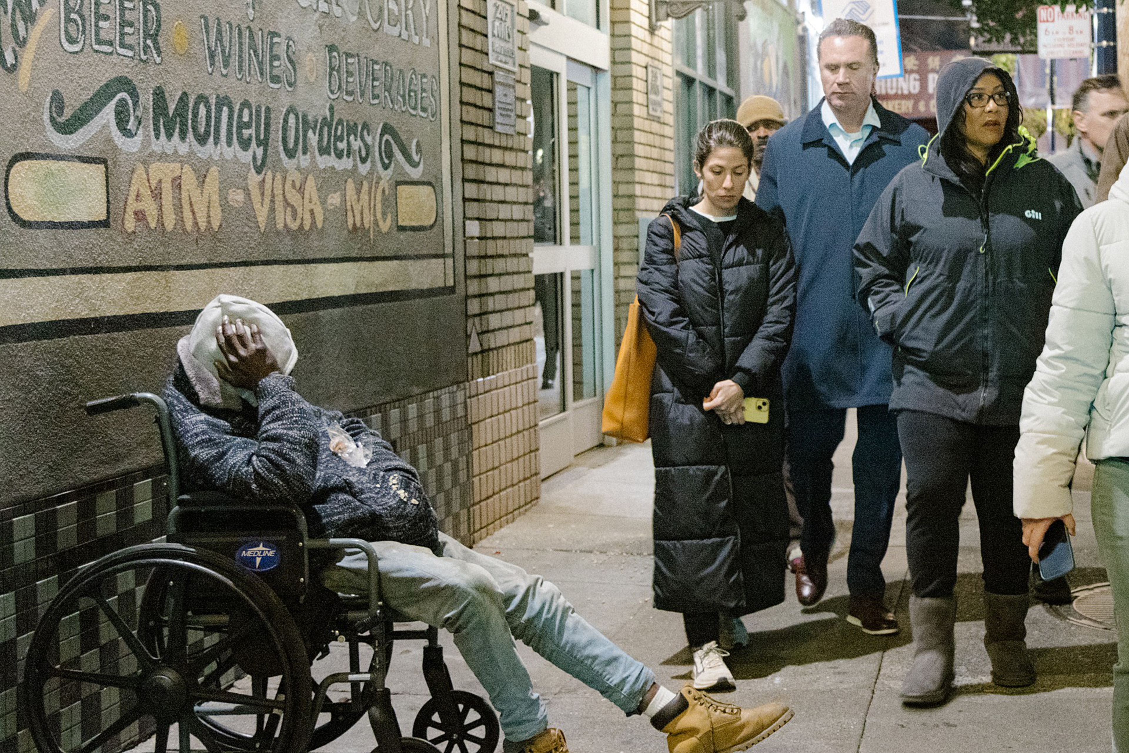 A person in a wheelchair, head in hands, sits by a wall as pedestrians walk by, seemingly unnoticed.