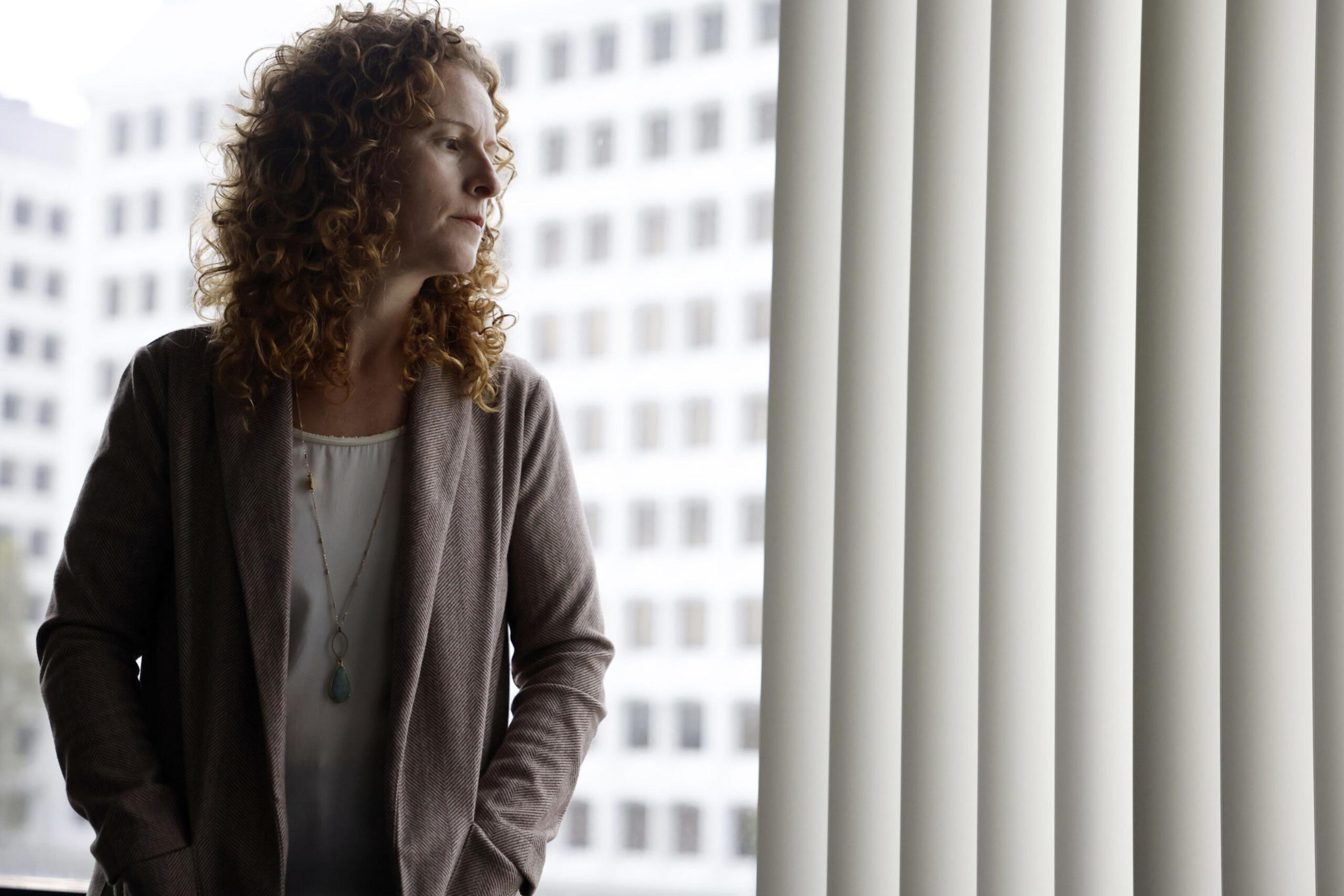 A woman with curly hair wearing a brown cardigan looks pensively out a window with vertical blinds in an office building setting.