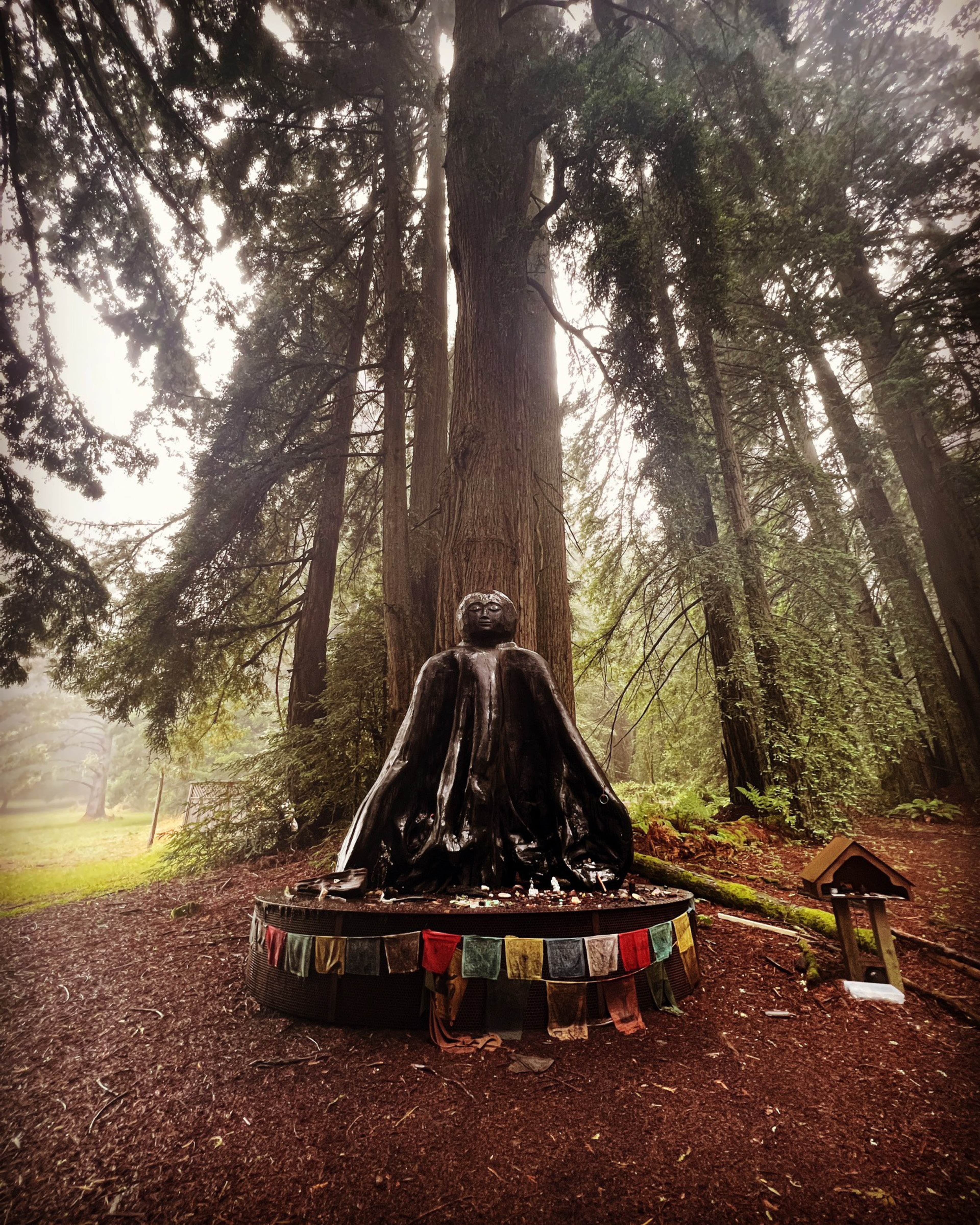 A dark statue of a seated figure draped in flowing robes sits on a platform adorned with colorful prayer flags, surrounded by tall, misty trees.