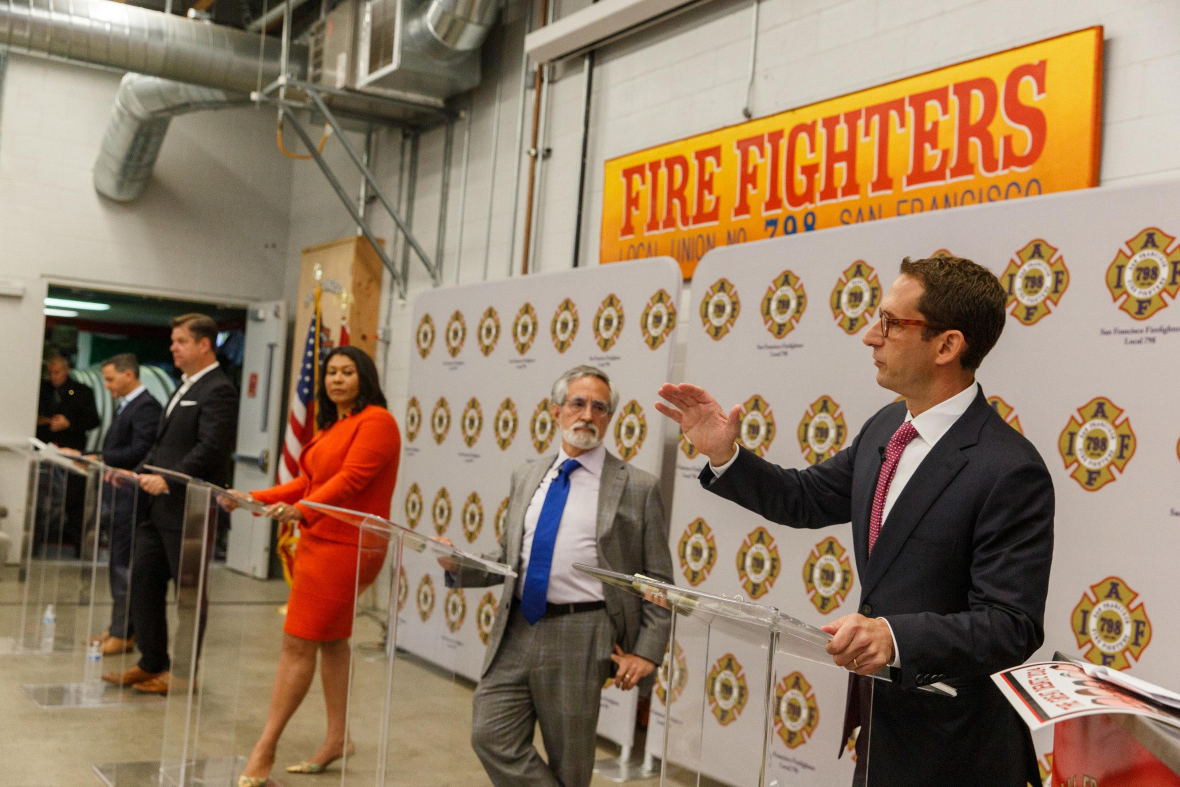 Four individuals stand behind clear podiums in a room with a yellow &quot;Fire Fighters&quot; sign and IAFF Local 798 logos, engaged in what appears to be a formal event or debate.