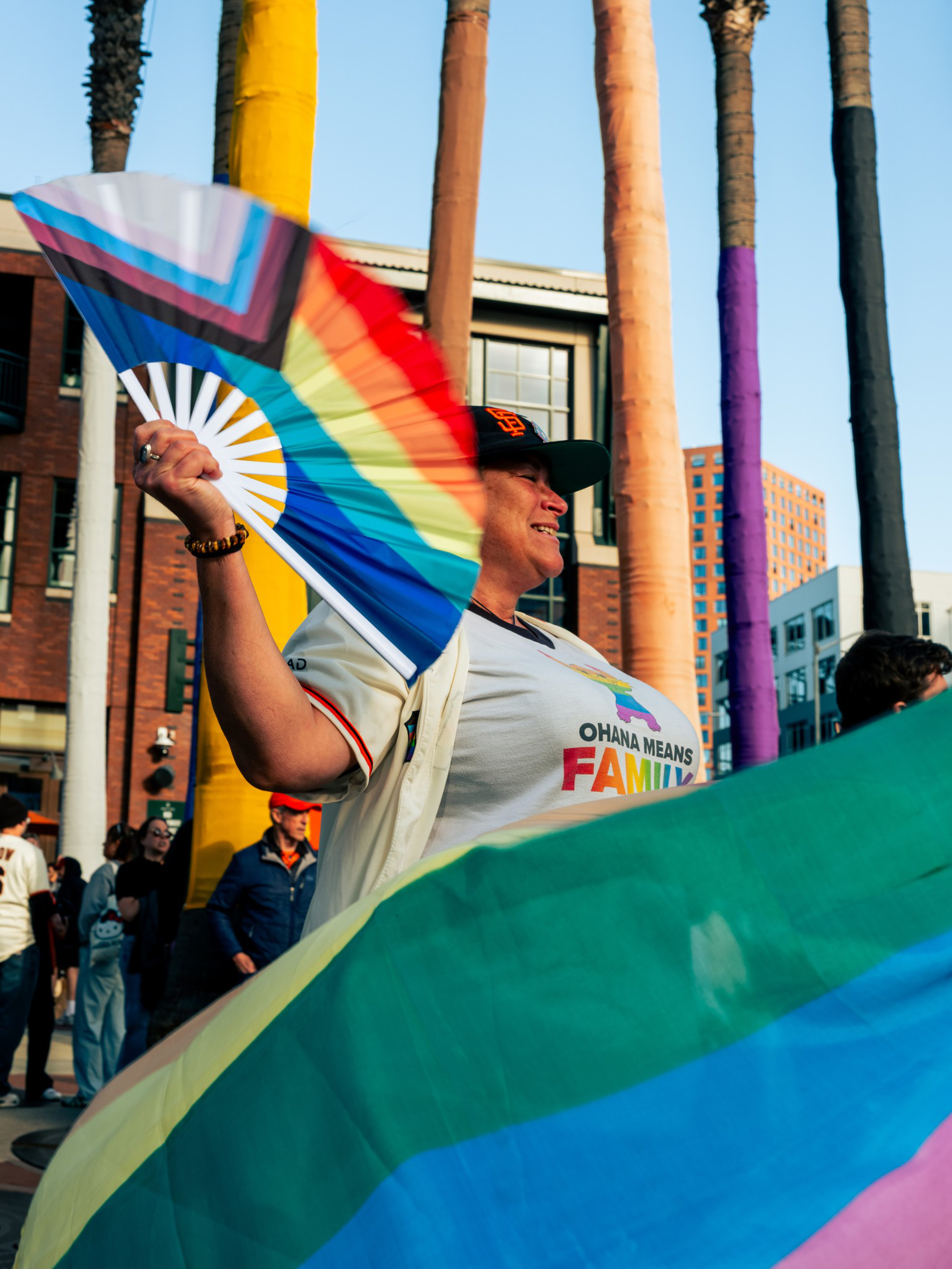 A person waves a rainbow pride fan and wears a shirt that says “Ohana means family,” with tall palm trees painted in various colors behind them.