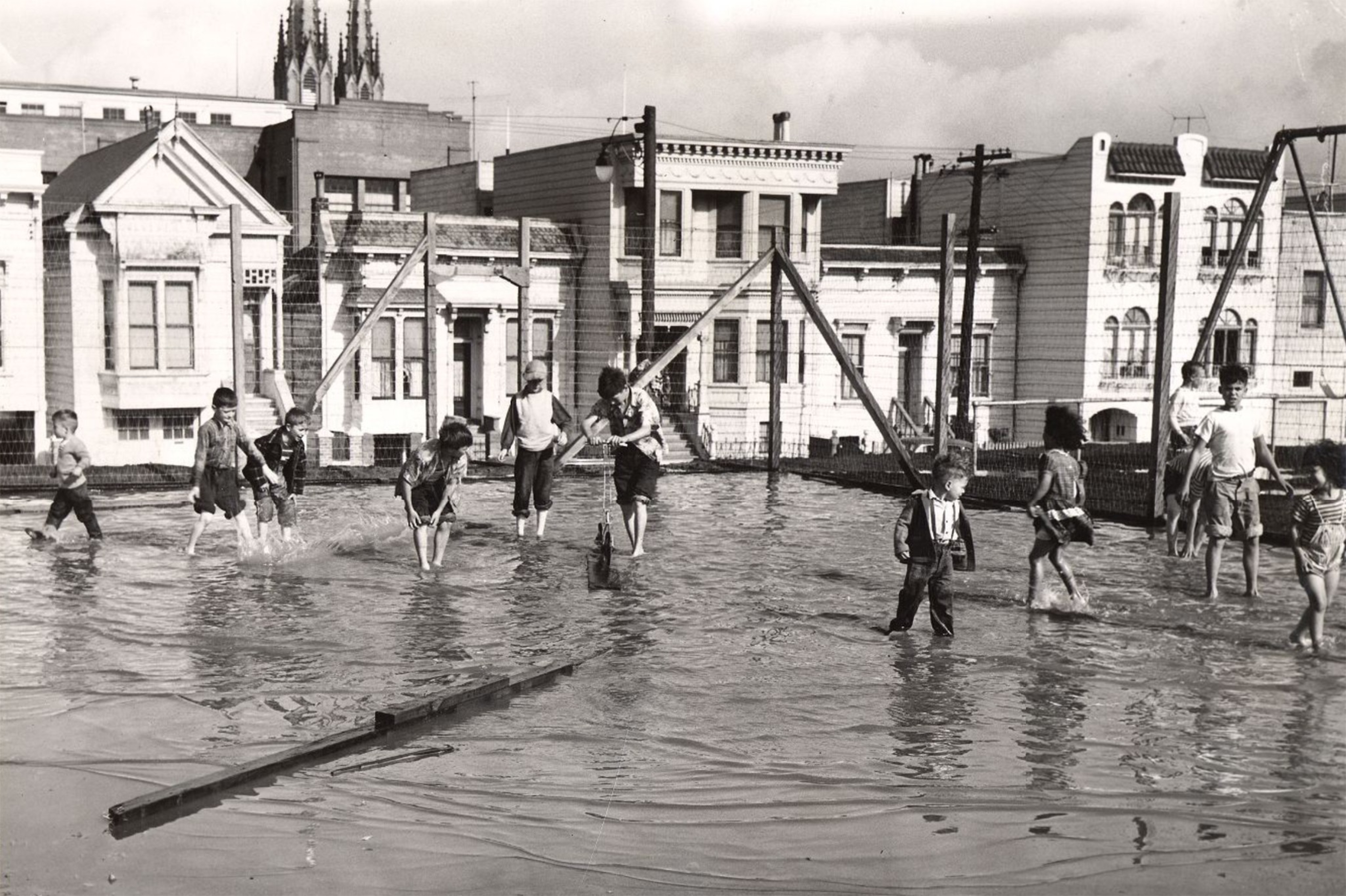 Children are playing and wading in a flooded playground with swings, surrounded by residential buildings in a black-and-white photo.