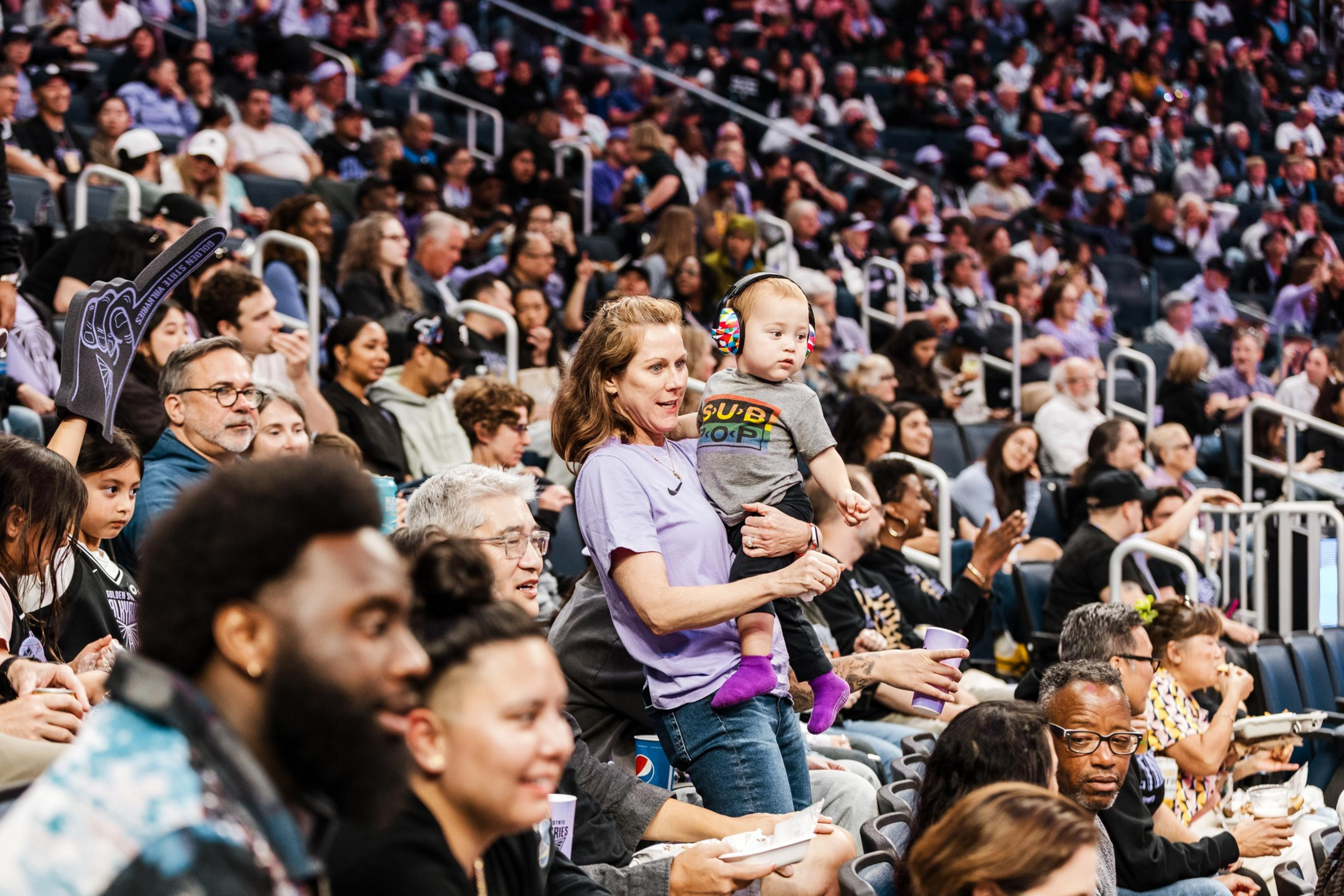 A woman holds a toddler wearing colorful earmuffs while a large crowd of diverse people watches an event in a stadium or arena.
