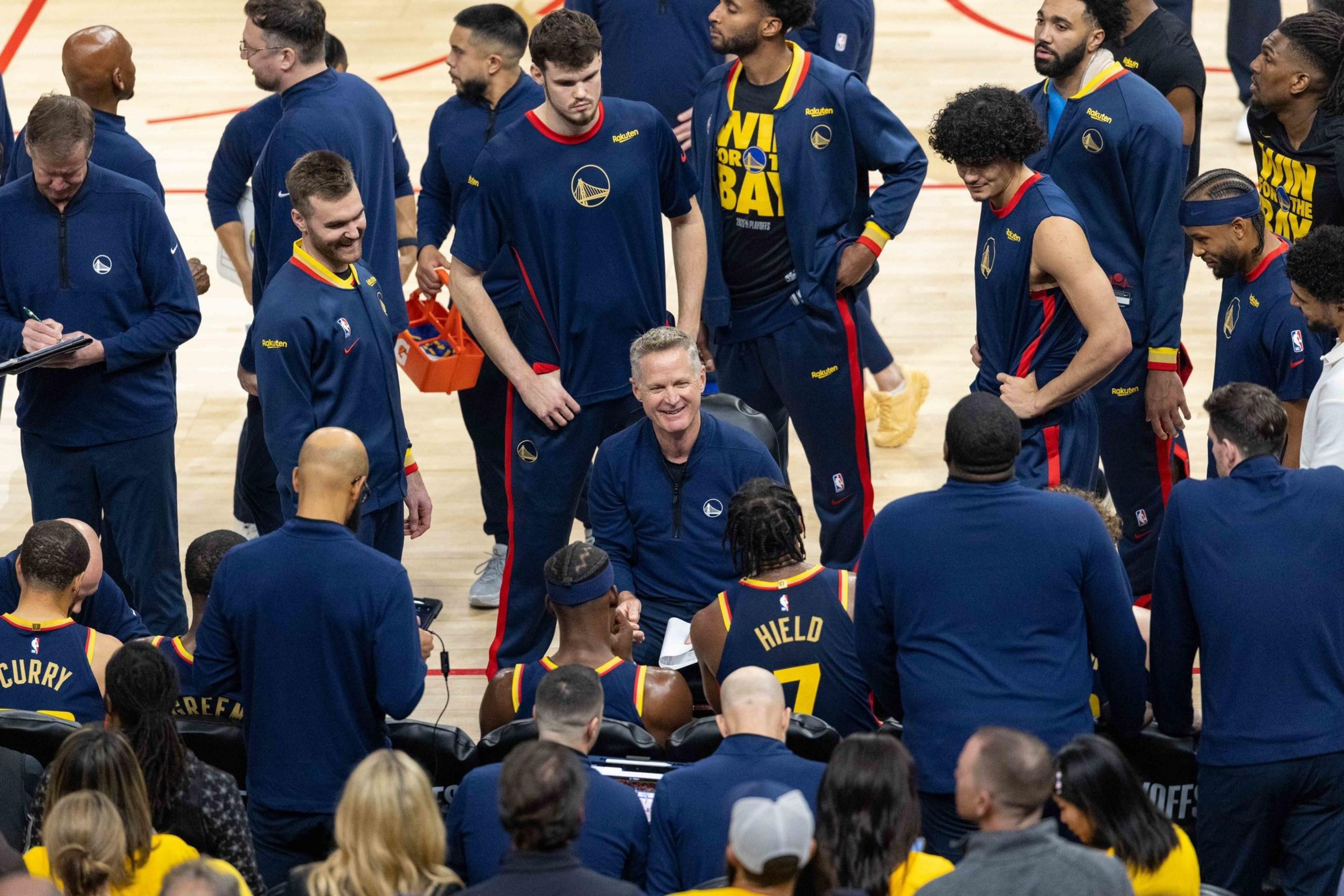 A basketball team huddles around a coach, who is smiling and seated, while players and staff stand around him wearing matching blue and yellow warm-up gear.