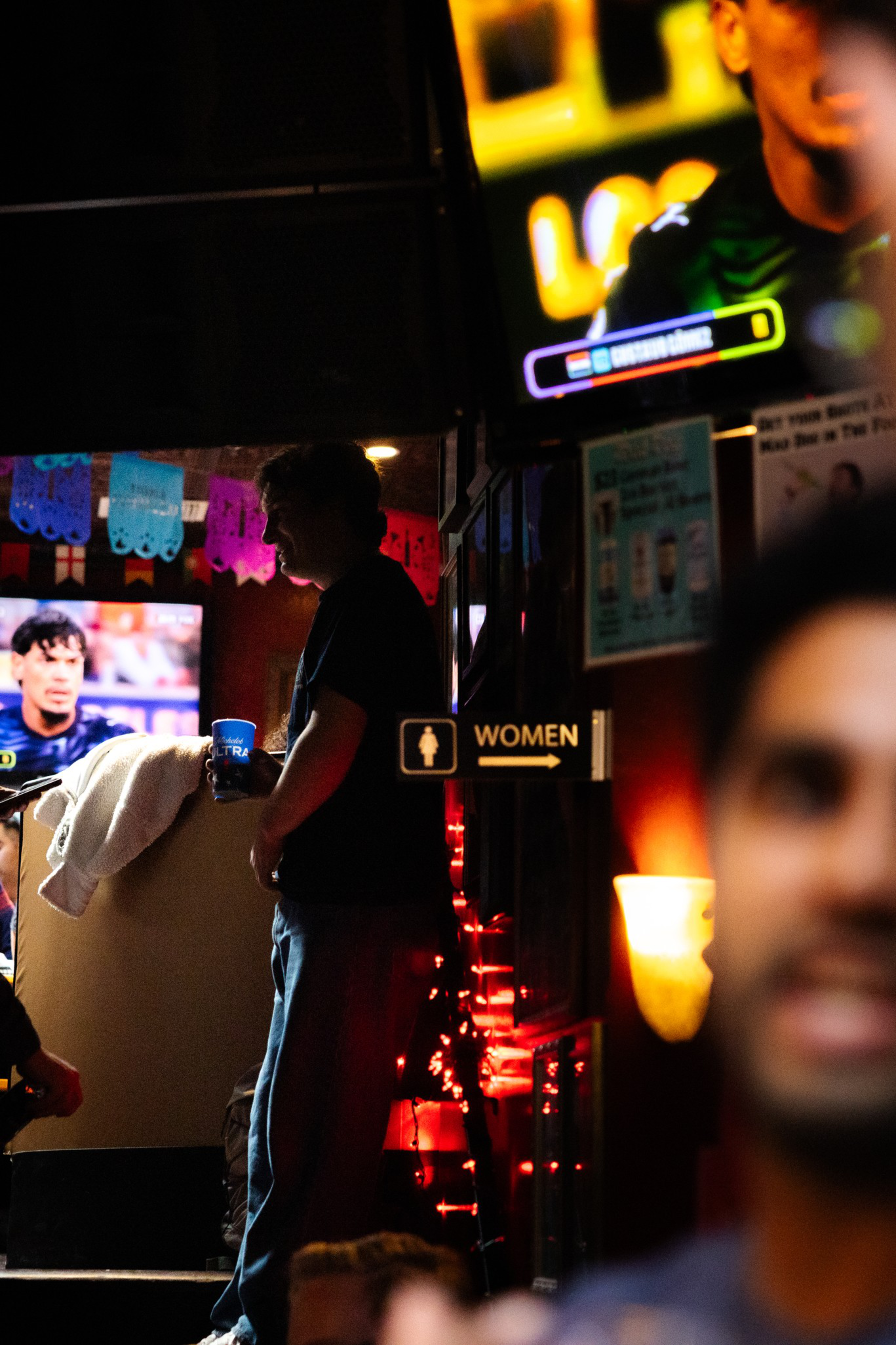 A dimly lit bar with a man holding a drink near a “WOMEN” restroom sign, colorful decorations, and two TVs showing a sports game.