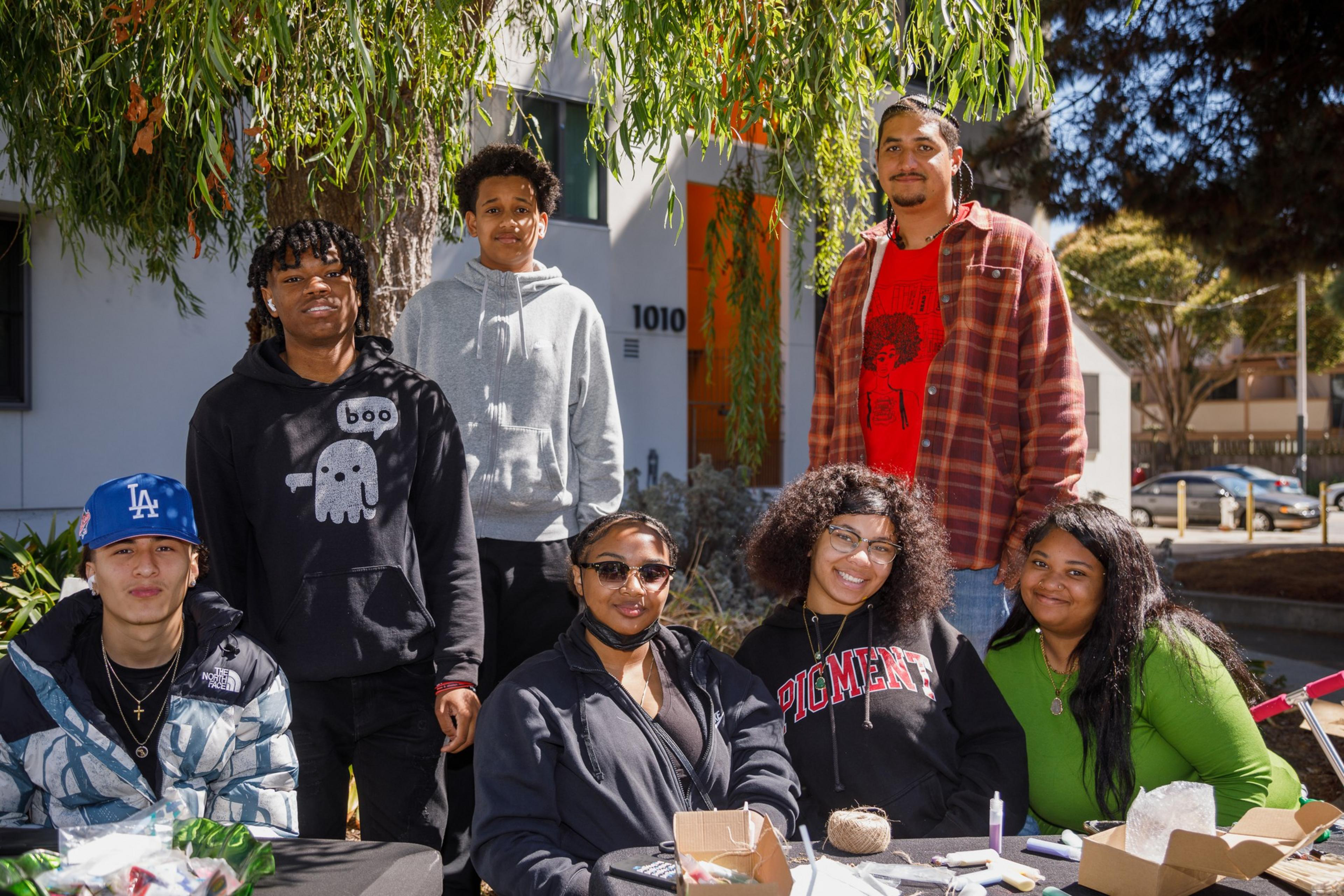 A group of seven young adults posing outdoors, some standing and some sitting around a table, with trees and a building in the background.