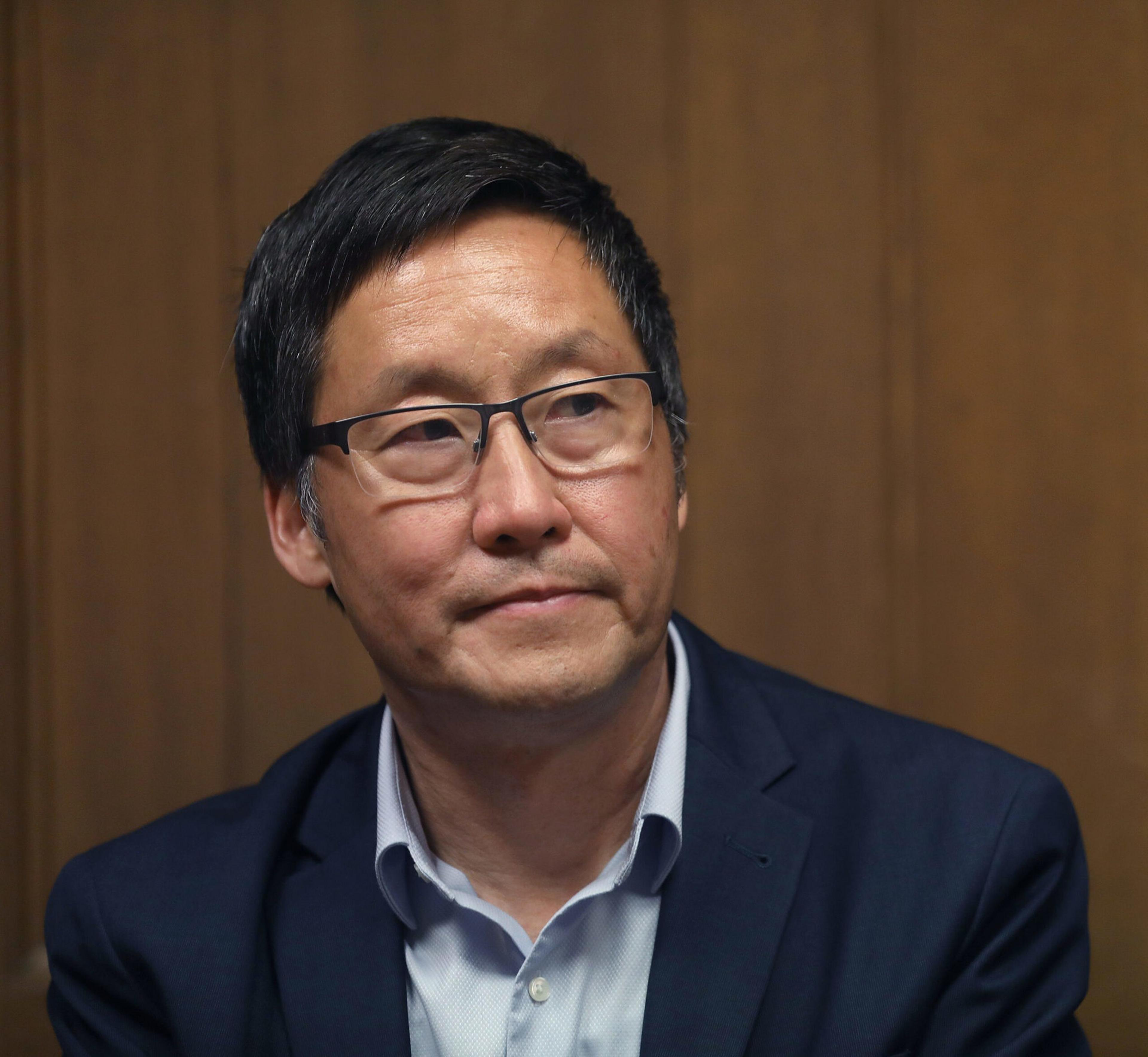 A middle-aged man with short black hair and glasses wears a blue suit jacket over a light blue shirt against a plain brown wooden background.