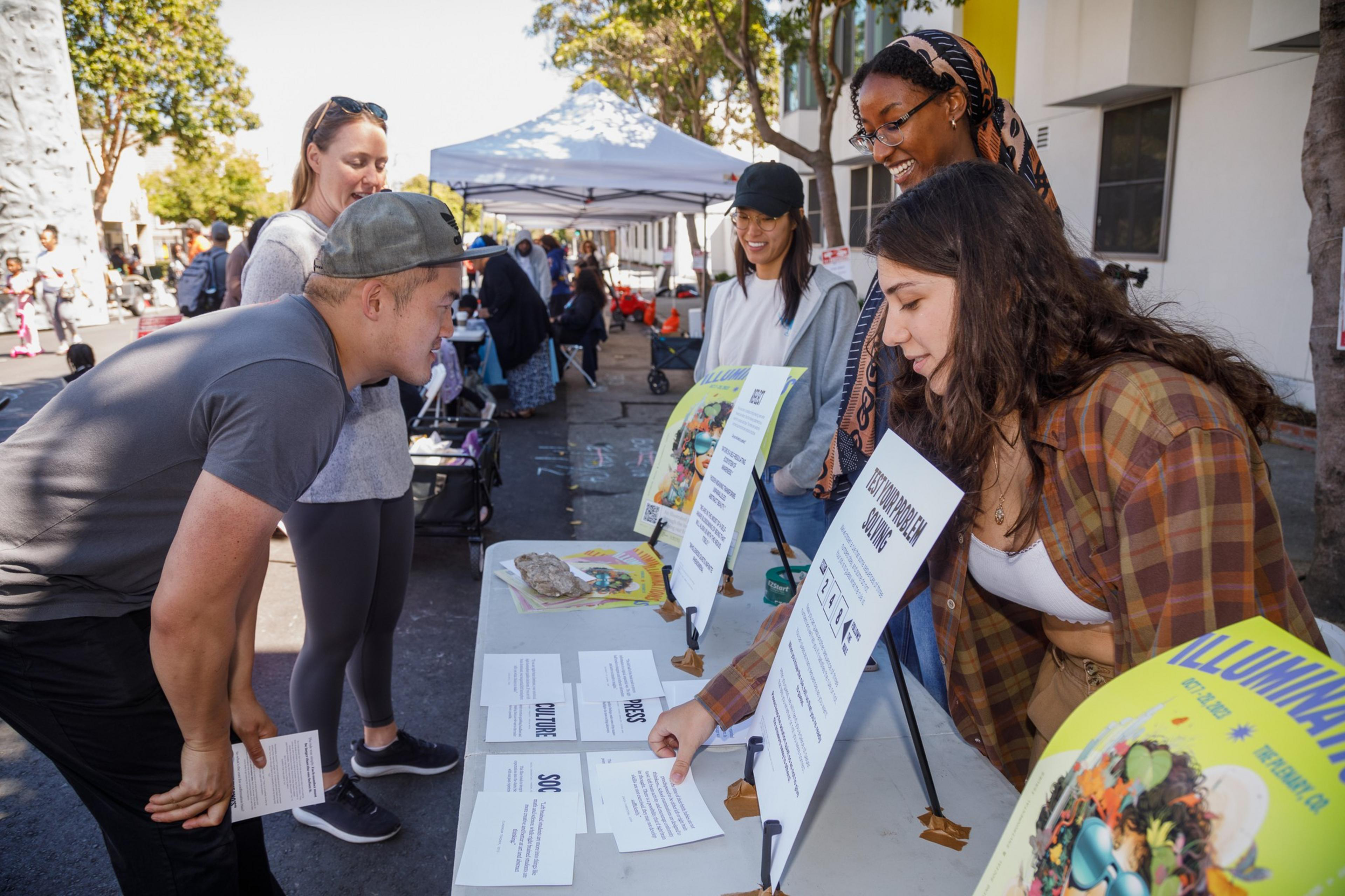 People are interacting at a sunny street fair, examining flyers and posters on a table.
